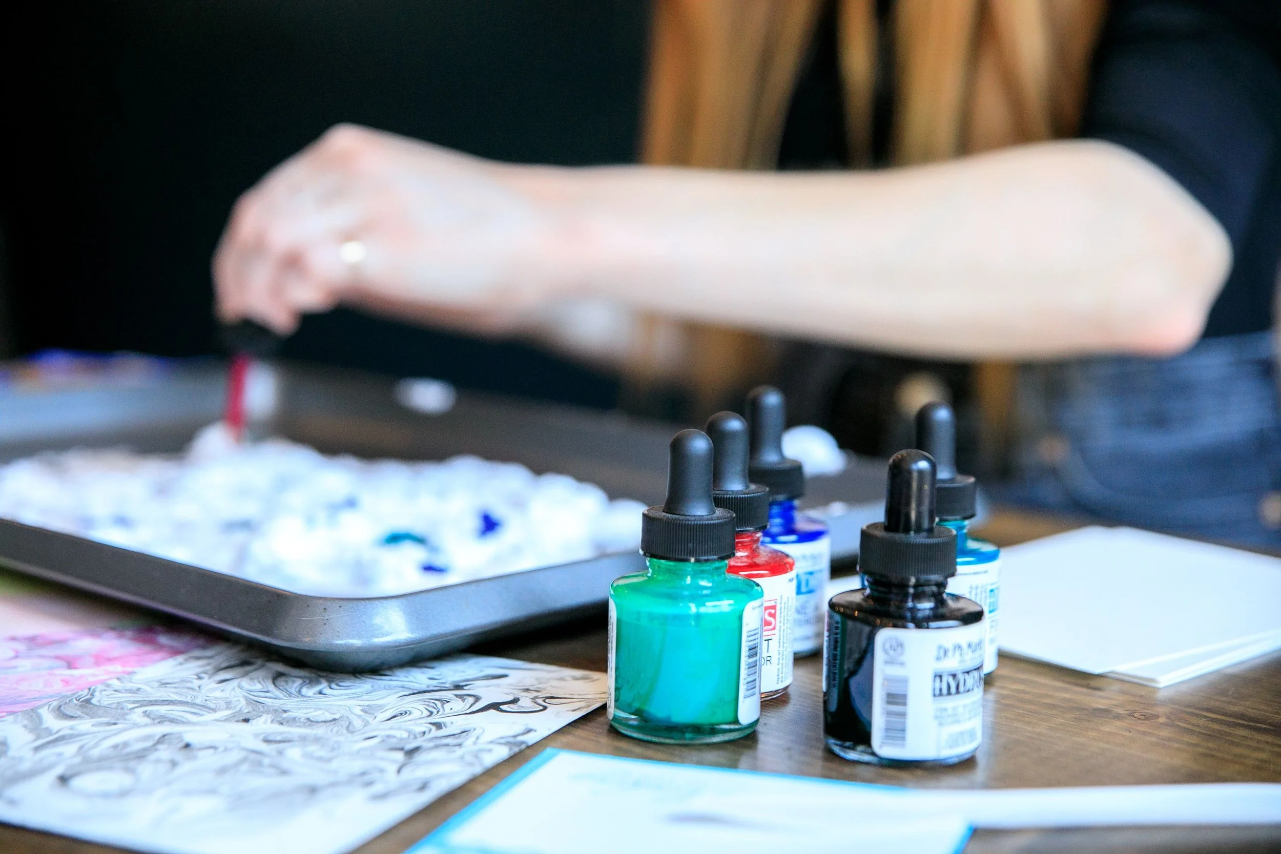 Close-up of four bottles of colorful paint on a table, with a splash of paint on paper and a blurred background of a person's arm and a mixing tray.