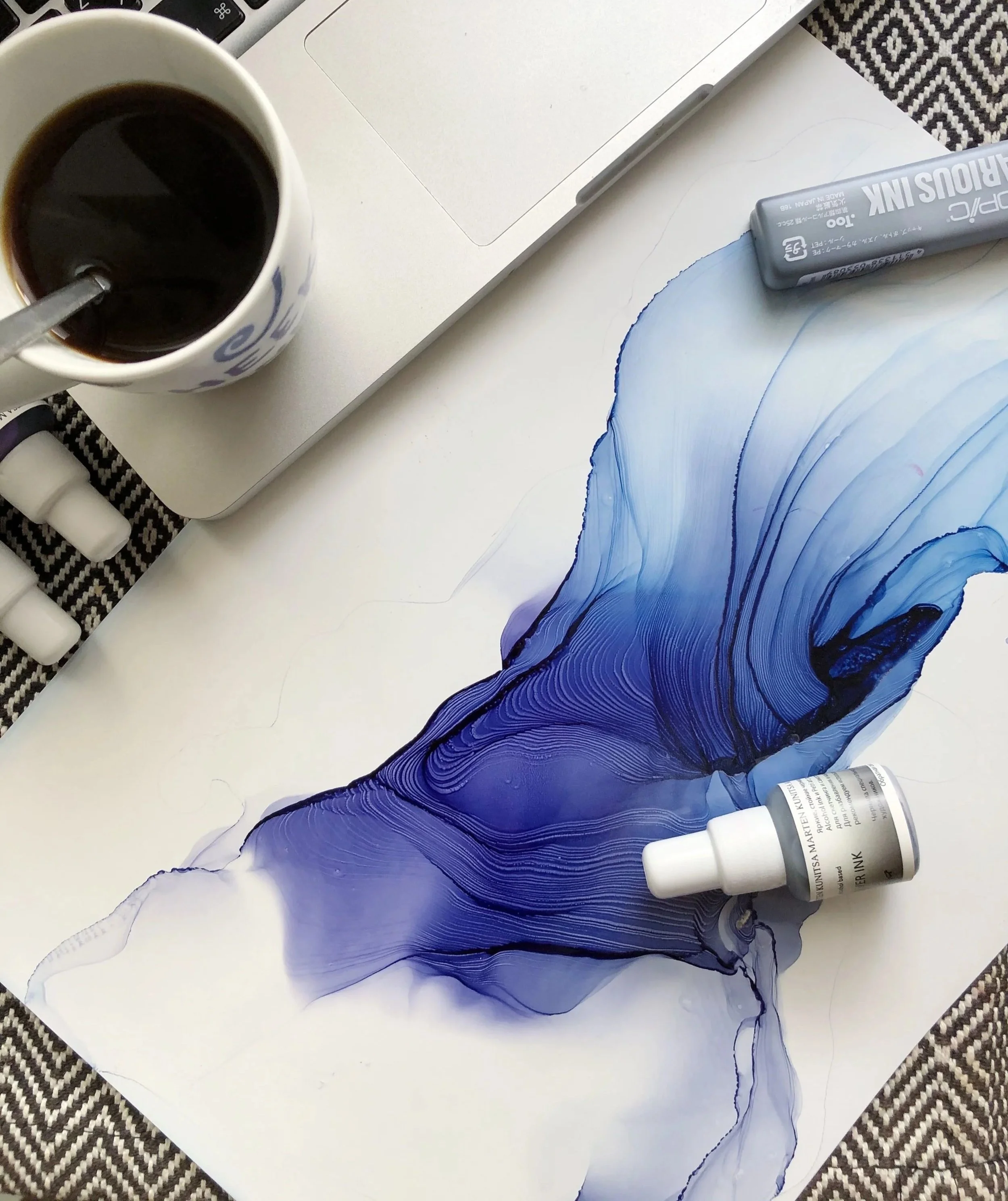 A white coffee mug filled with black coffee and a spoon resting inside, a silver laptop, a tube of alcohol ink, and a blue and black abstract watercolor painting on a white table with a black and white patterned cloth underneath.