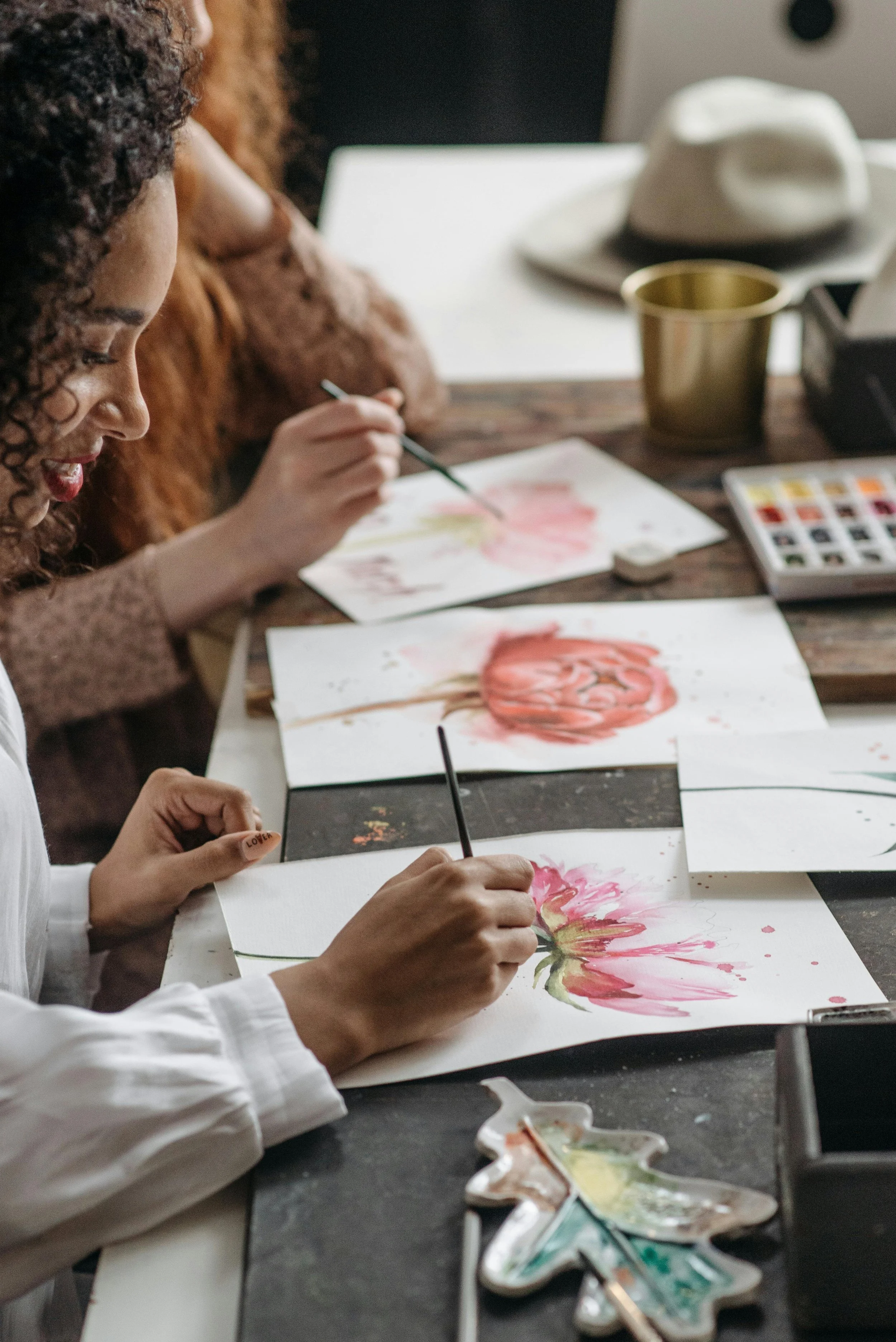 People are painting and creating watercolor floral artwork at a table, surrounded by art supplies and cups.