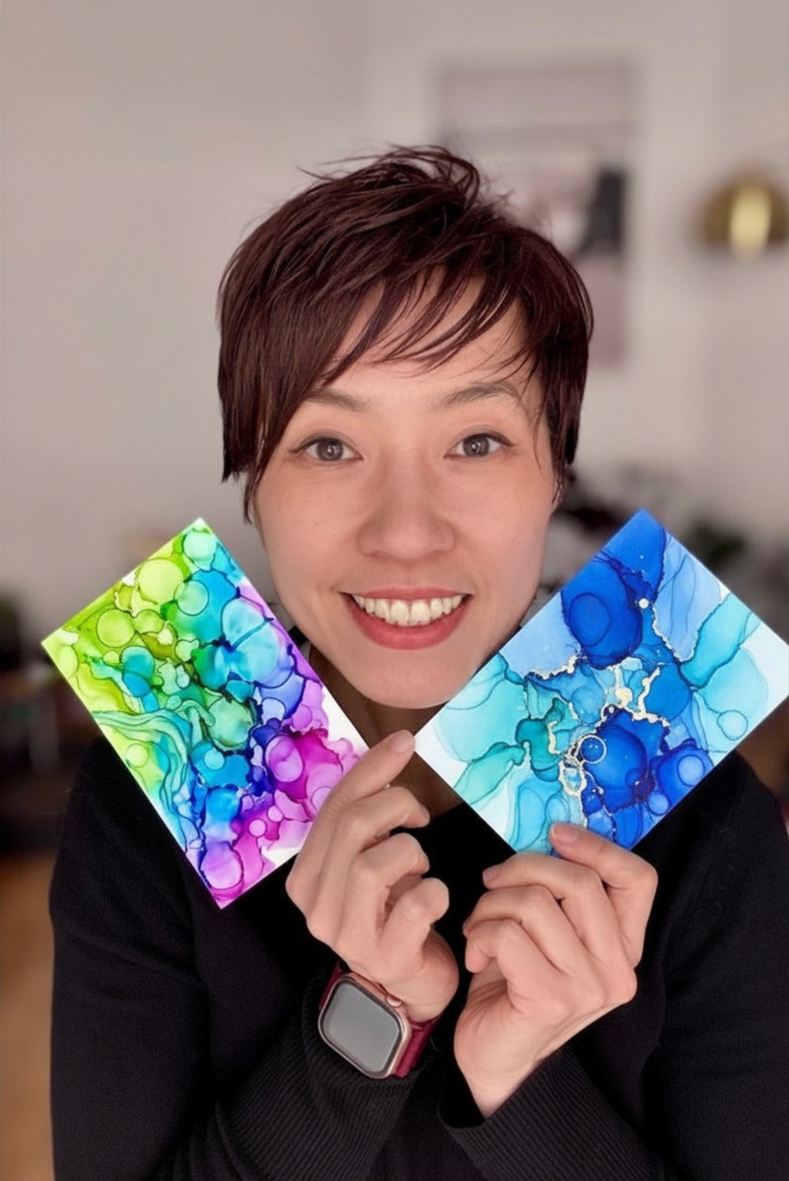 A woman with short brown hair smiling and holding three colorful abstract paintings, one green and purple, one blue and white, and one pink, blue, and purple.