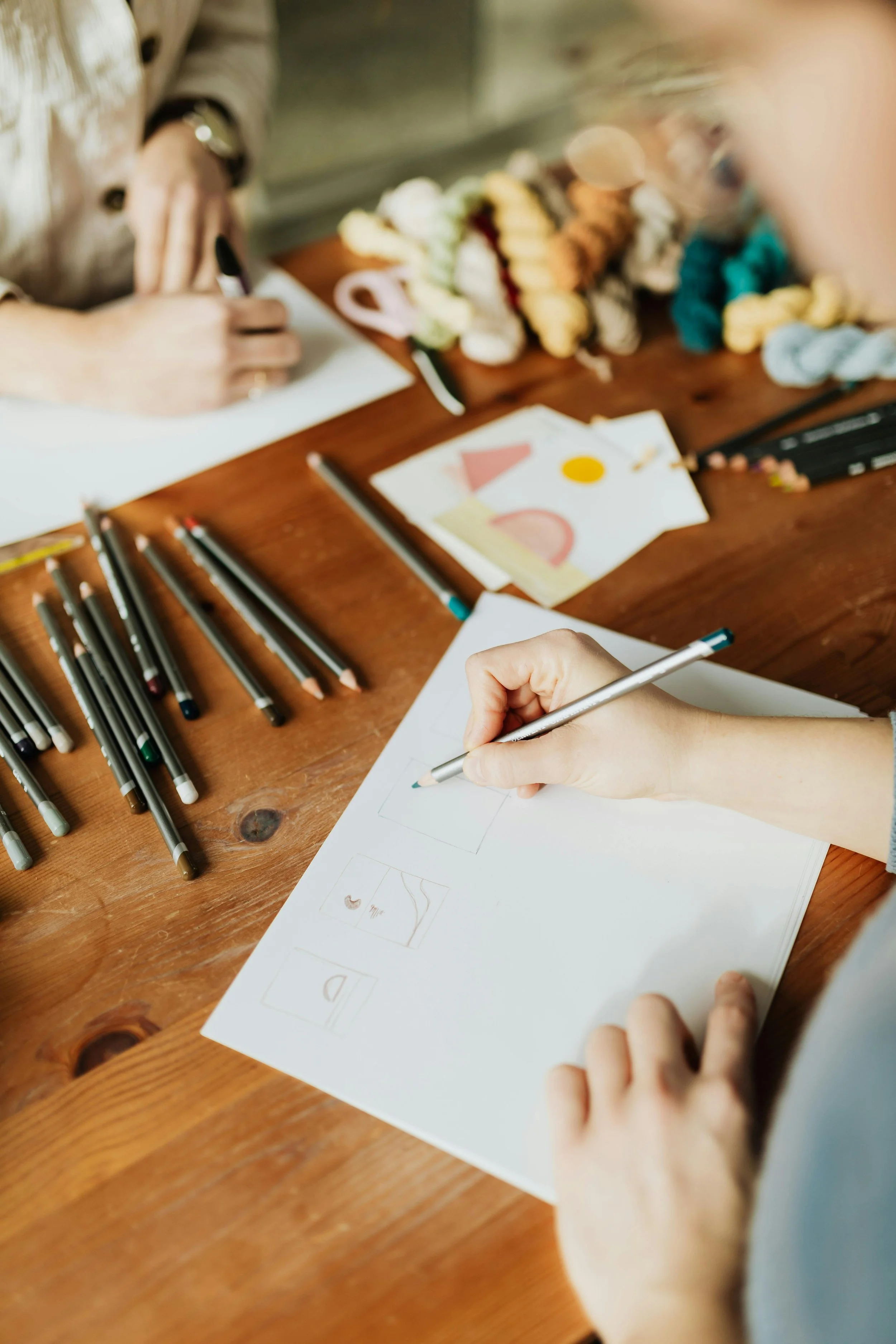 Person drawing and coloring on paper, surrounded by markers, yarn, and other art supplies on a wooden table.