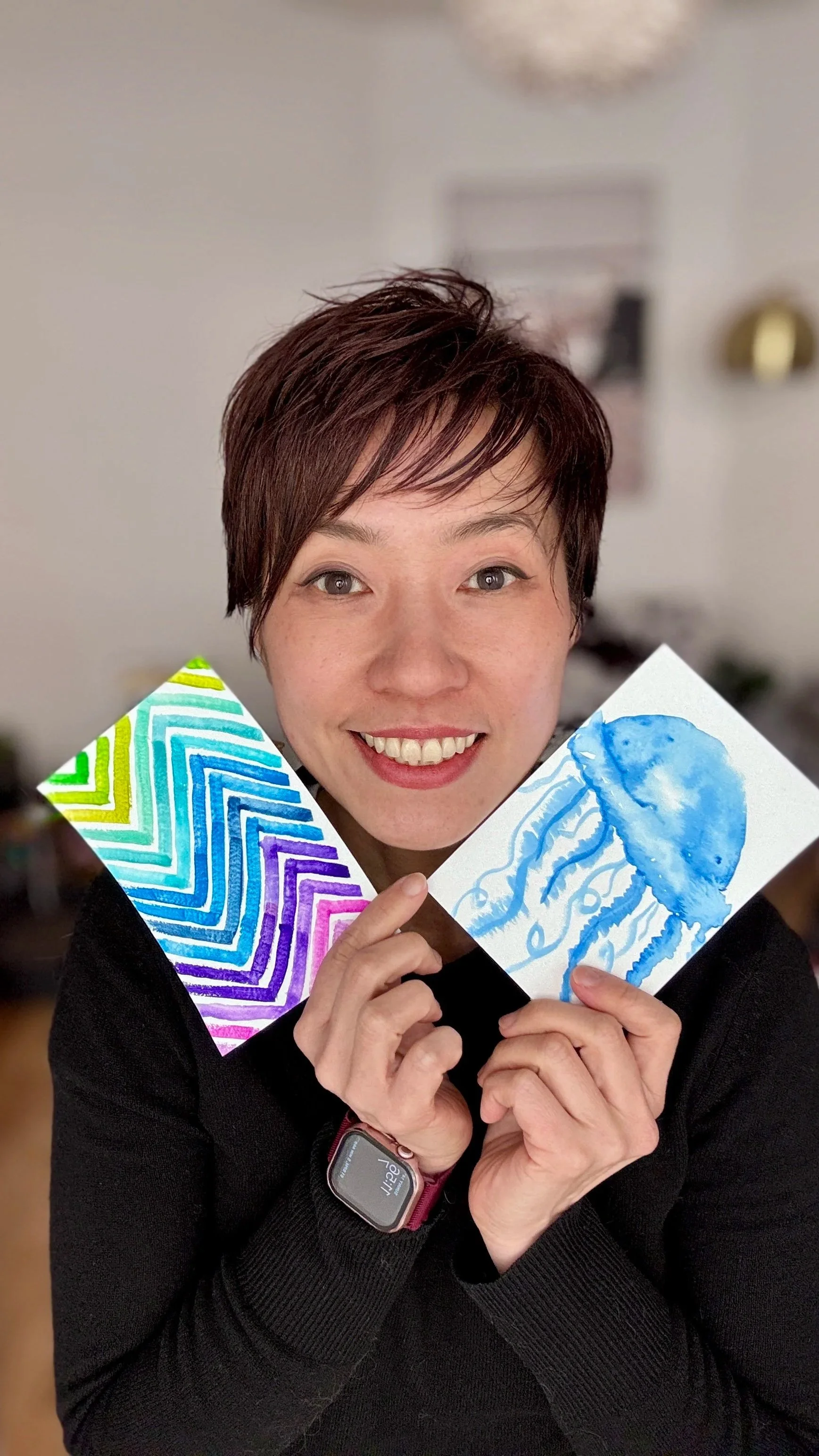 A woman with short brown hair smiling and holding two colorful paintings, one with rainbow-colored chevron pattern and another with blue watercolor jellyfish, in an indoor setting.