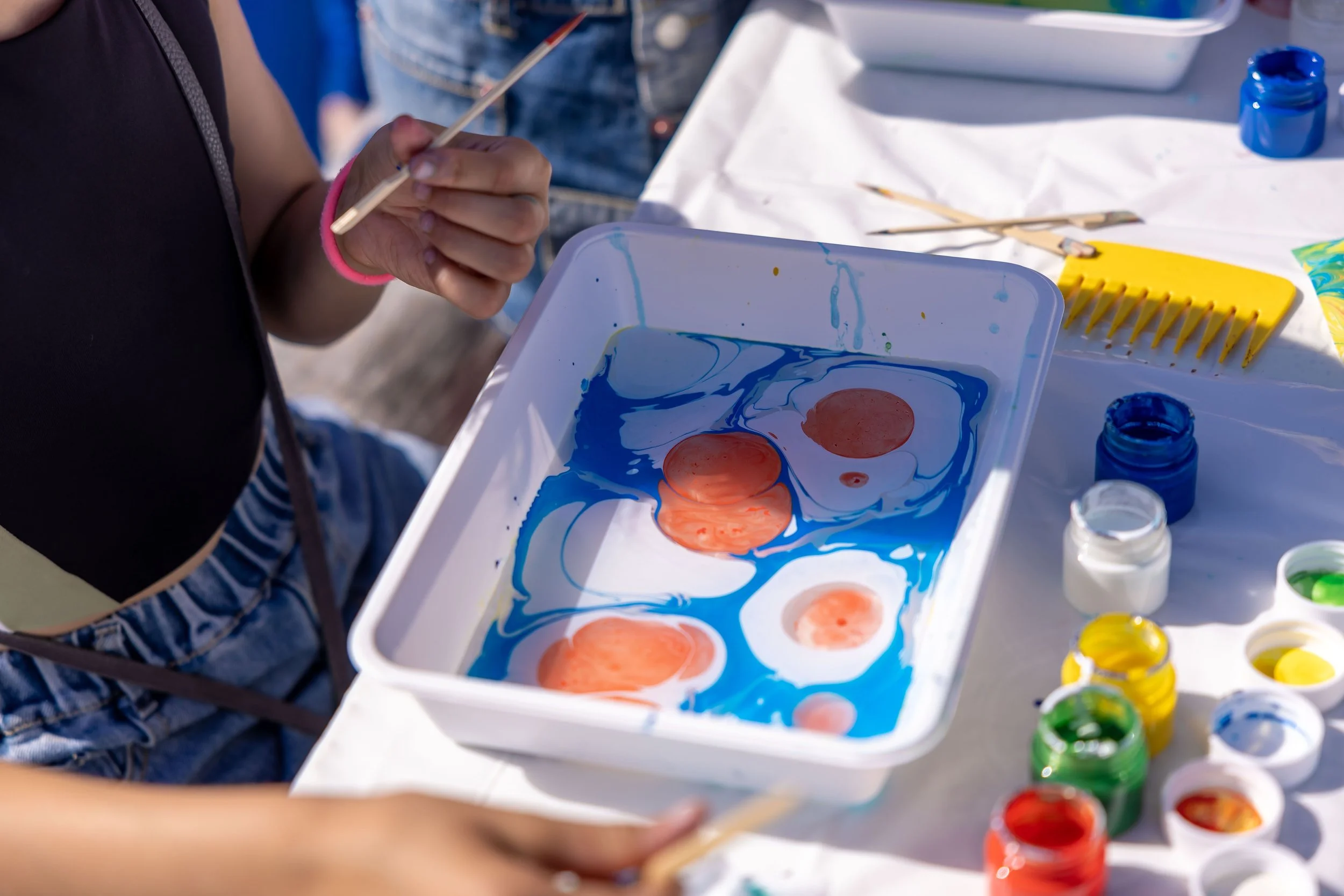 Person holding a paintbrush near a palette with orange lipstick-shaped paint blobs ready for ebru marbling, surrounded by open bottles of blue, green, yellow, and white paint, with other painting tools on a white table.