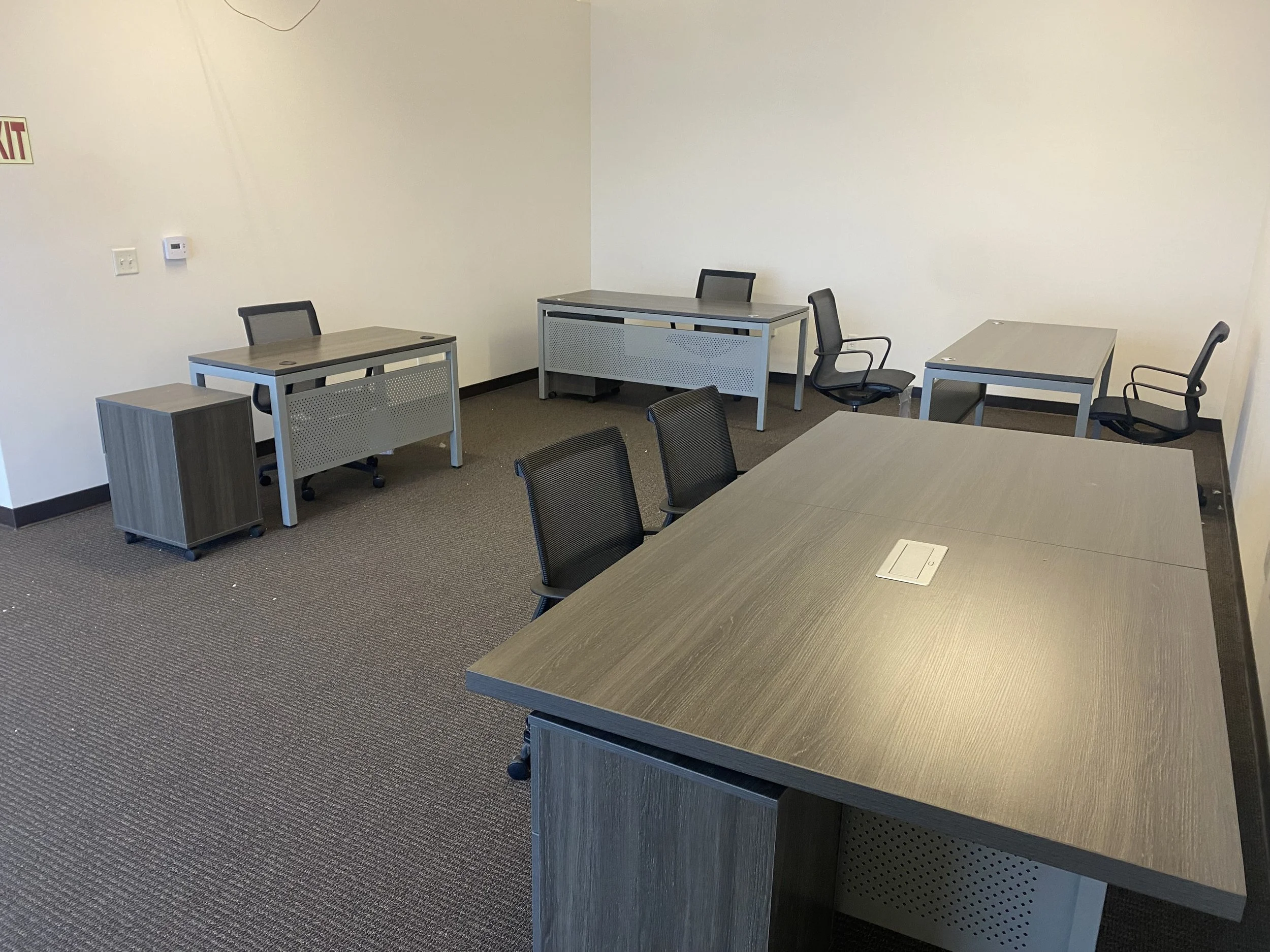 Empty office room with four desks and six chairs, beige walls, gray carpet, and an exit sign.