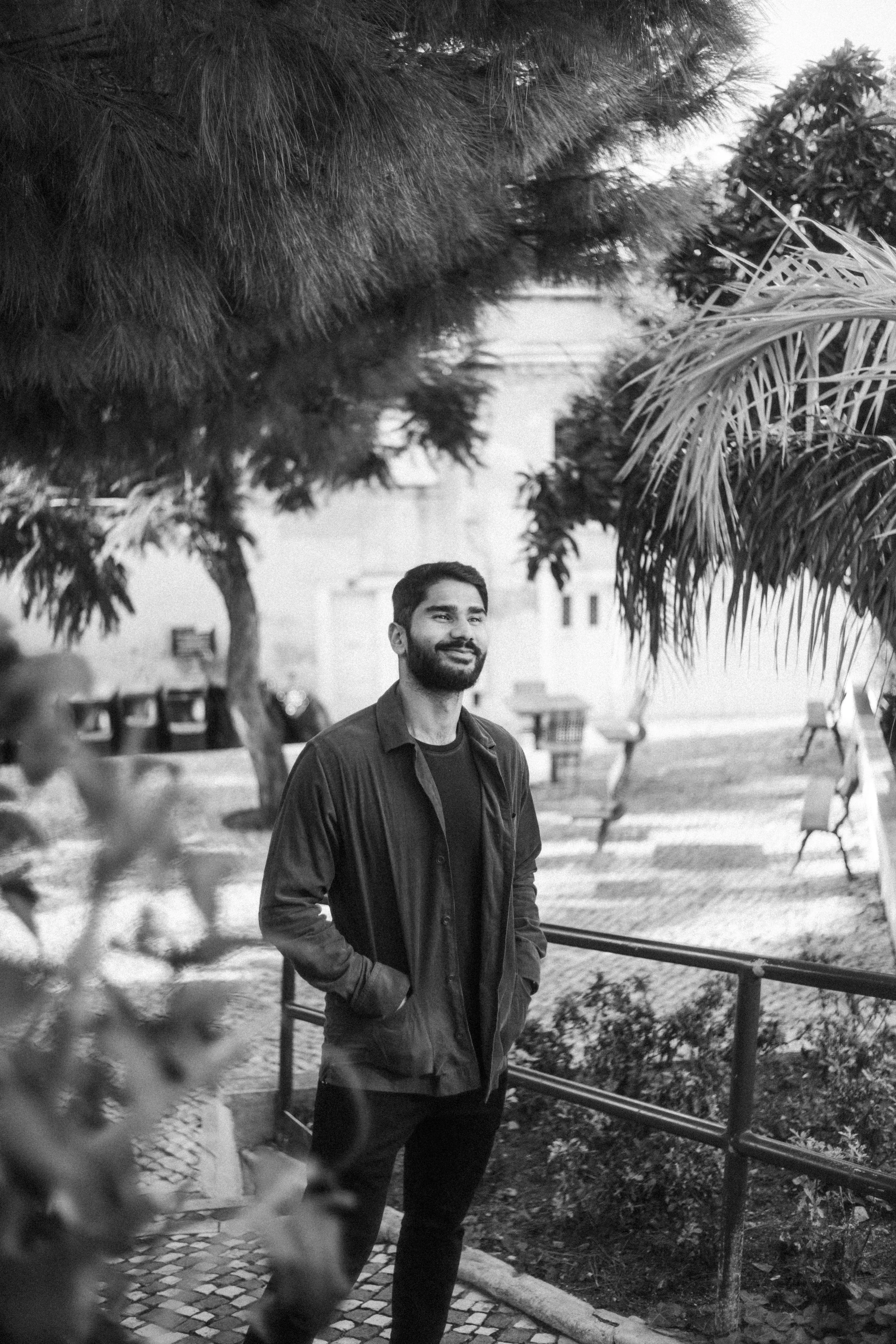 A man with a beard standing outdoors with trees and benches, hands in his jacket pockets, smiling in black and white.