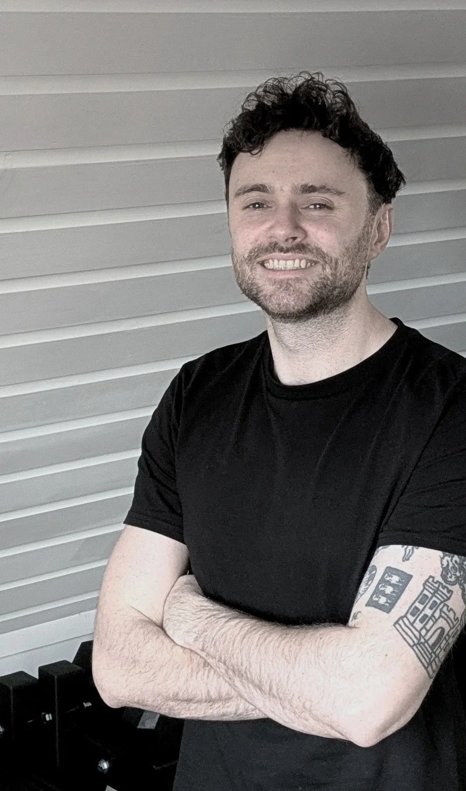 A man with curly dark hair and a beard is smiling with arms crossed, wearing a black t-shirt, standing in front of a horizontal white wall.