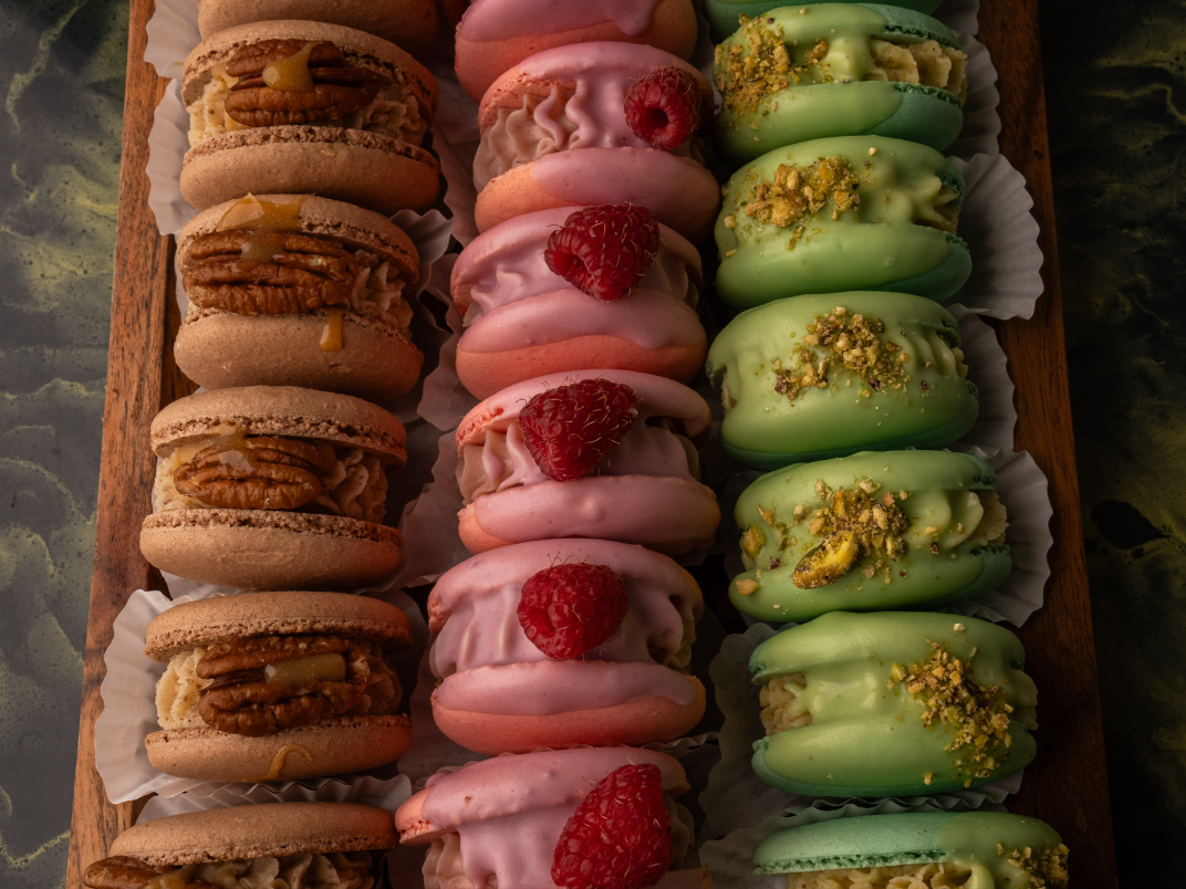 Display of three rows of assorted macarons on a wooden tray, including caramel pecan, raspberry, and pistachio flavors.