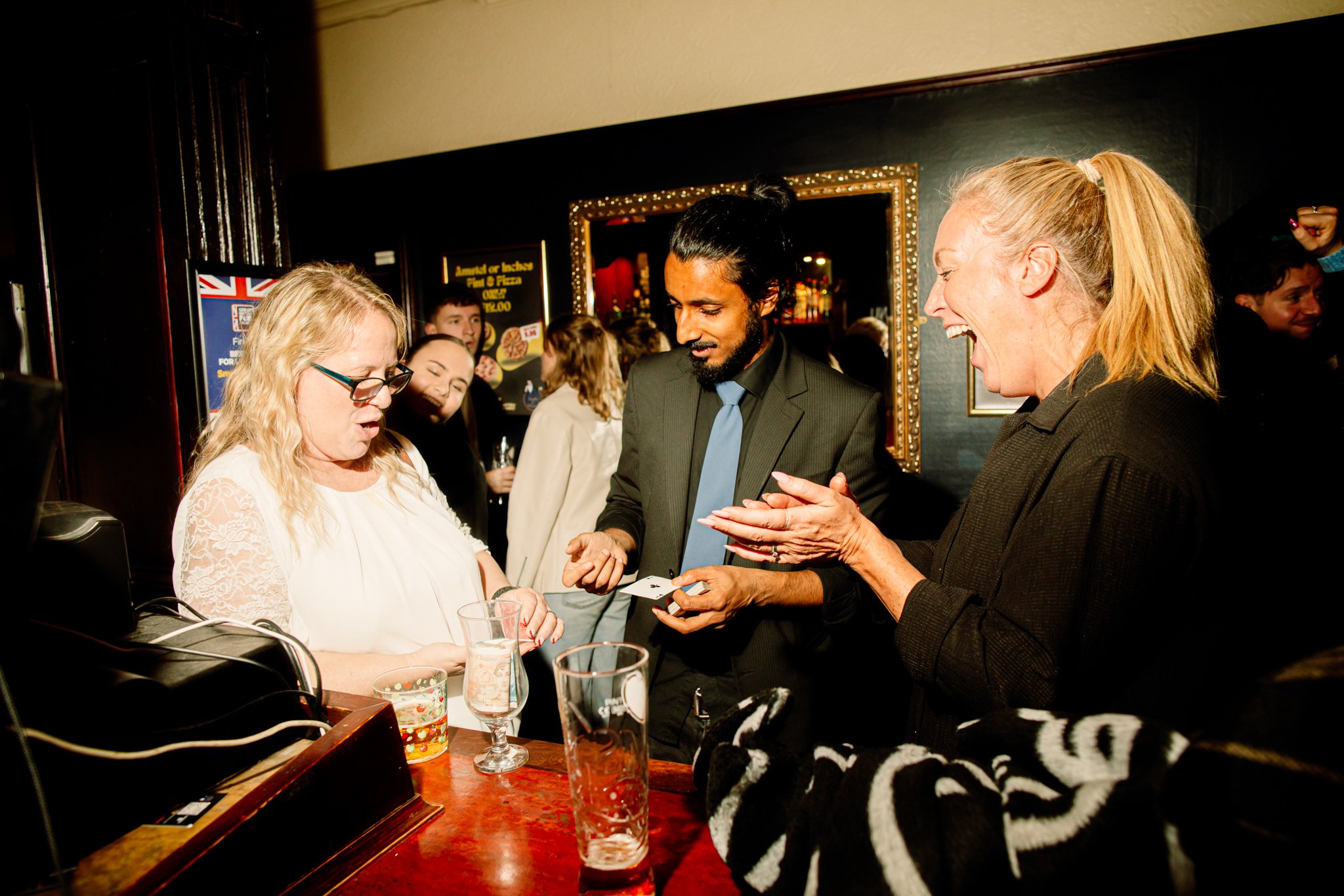 A woman with glasses looks surprised as a magician performs a card trick at a bar, while another woman laughs in excitement, and several people are gathered in the background.