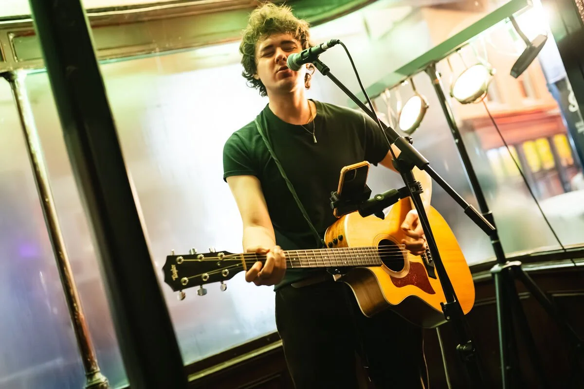 Young person singing into a microphone while playing an acoustic guitar, with stage lighting and equipment in the background.