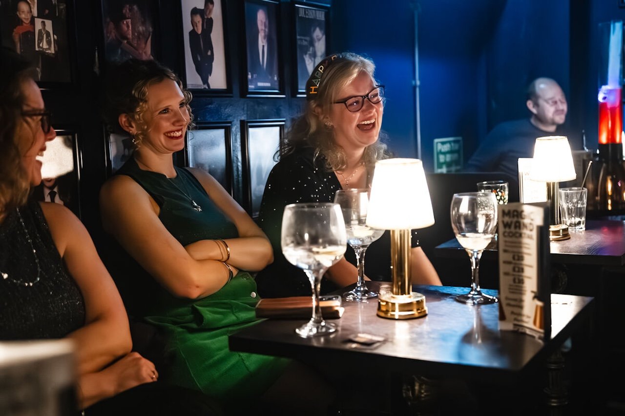 People sitting at a bar table, smiling and laughing, with drinks and lamps, in a dimly lit bar or pub.