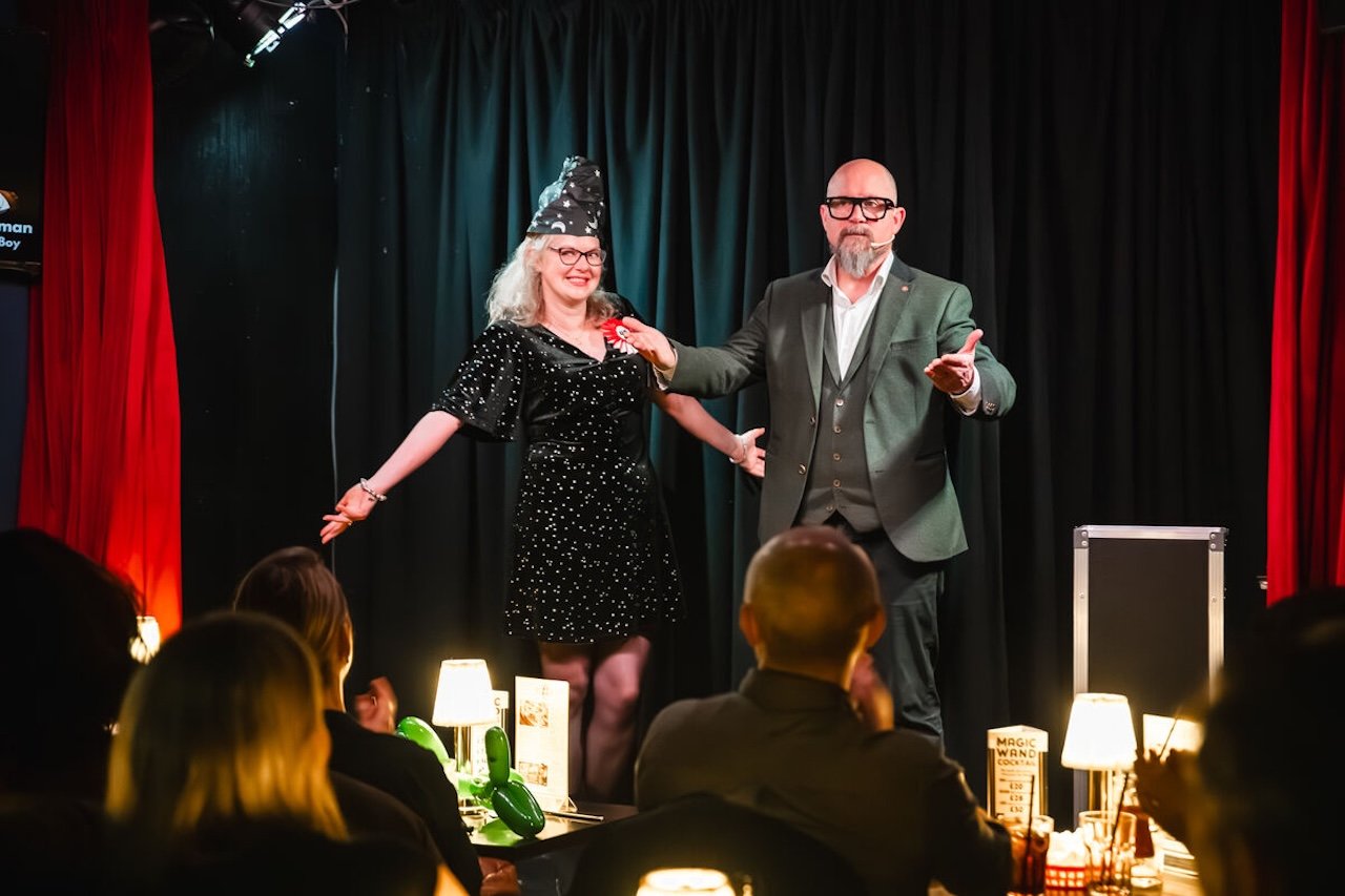A woman in a black dress with white specks and a tall black hat stands on stage with a man in a gray suit. The woman is smiling, and the man is gesturing with his hand. The stage has black curtains with red side drapes, and audience members are seated at tables with lamps and drinks, watching the performance.