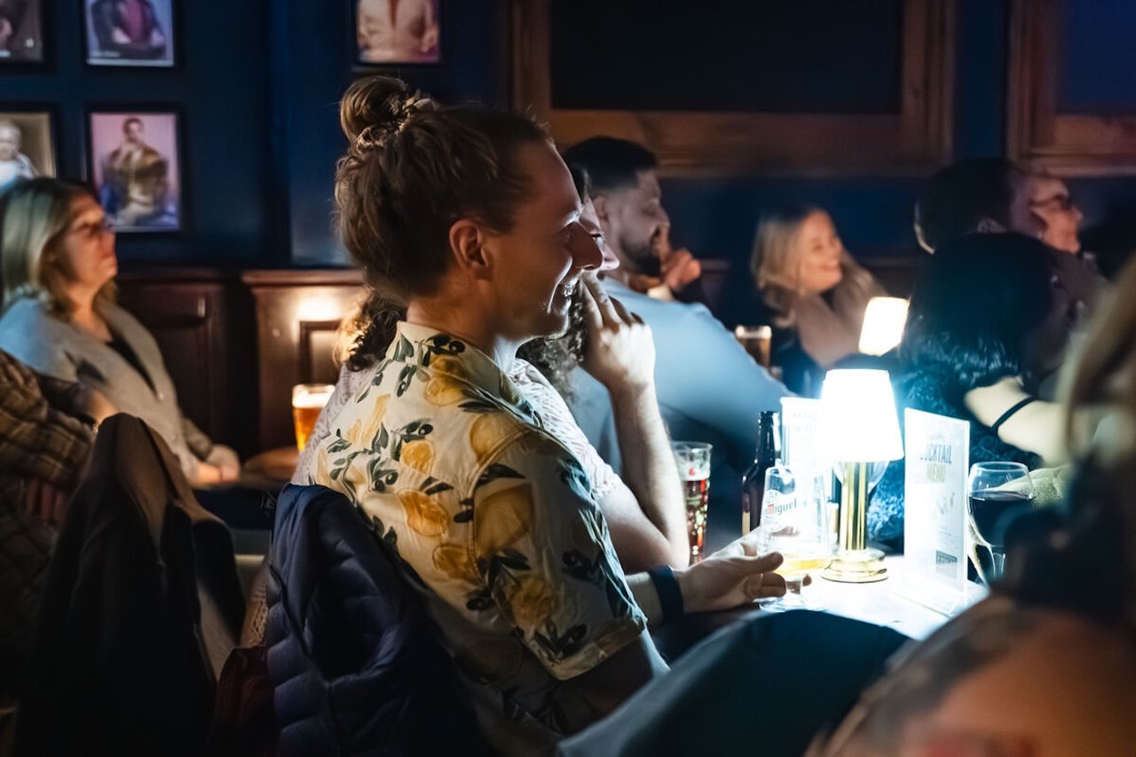 A woman with a floral shirt sitting at a bar or pub, smiling and looking towards the front. Several other people are seated around her, some with drinks, in a dimly-lit setting, possibly during a comedy or karaoke night.
