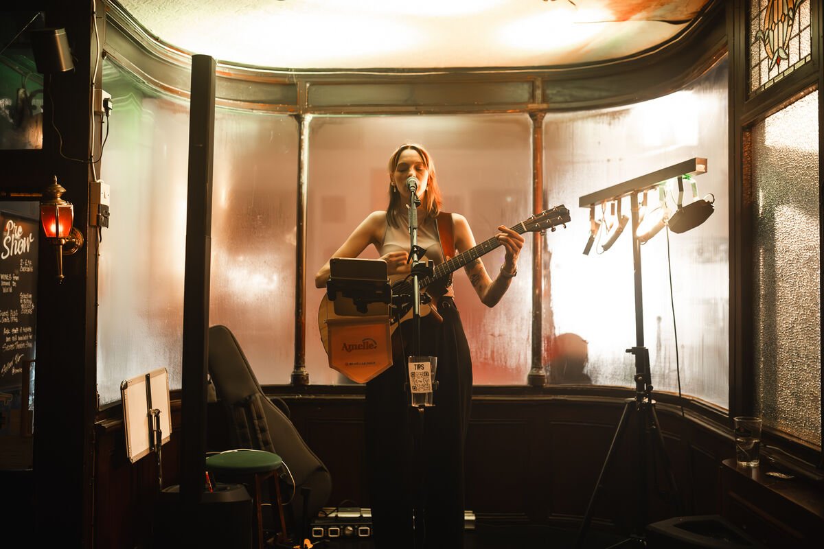 A woman performing with an acoustic guitar and microphone on a small stage in a dimly lit venue.