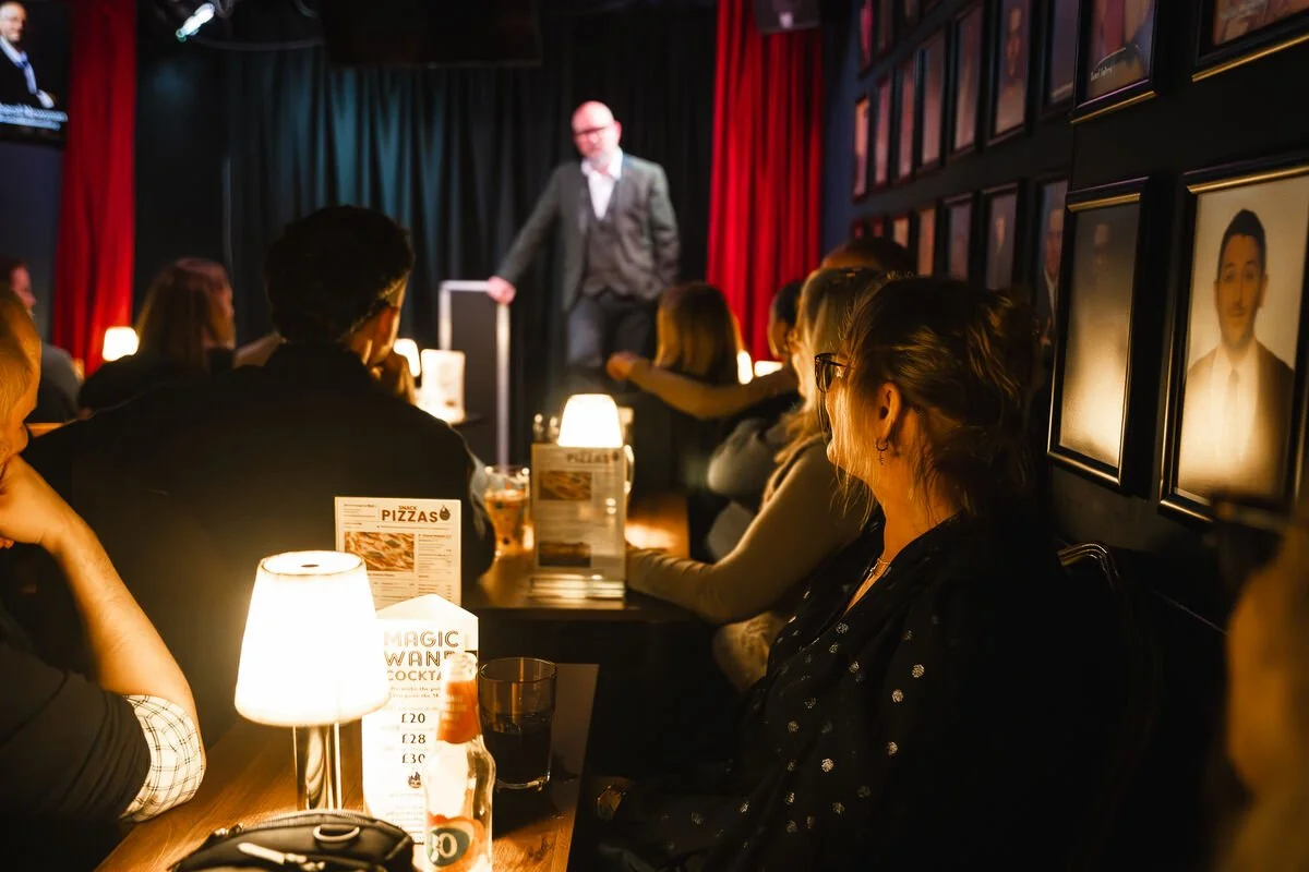 A comedy club with people seated at tables watching a comedian perform on stage. The venue has dim lighting, framed pictures on the wall, and small table lamps.