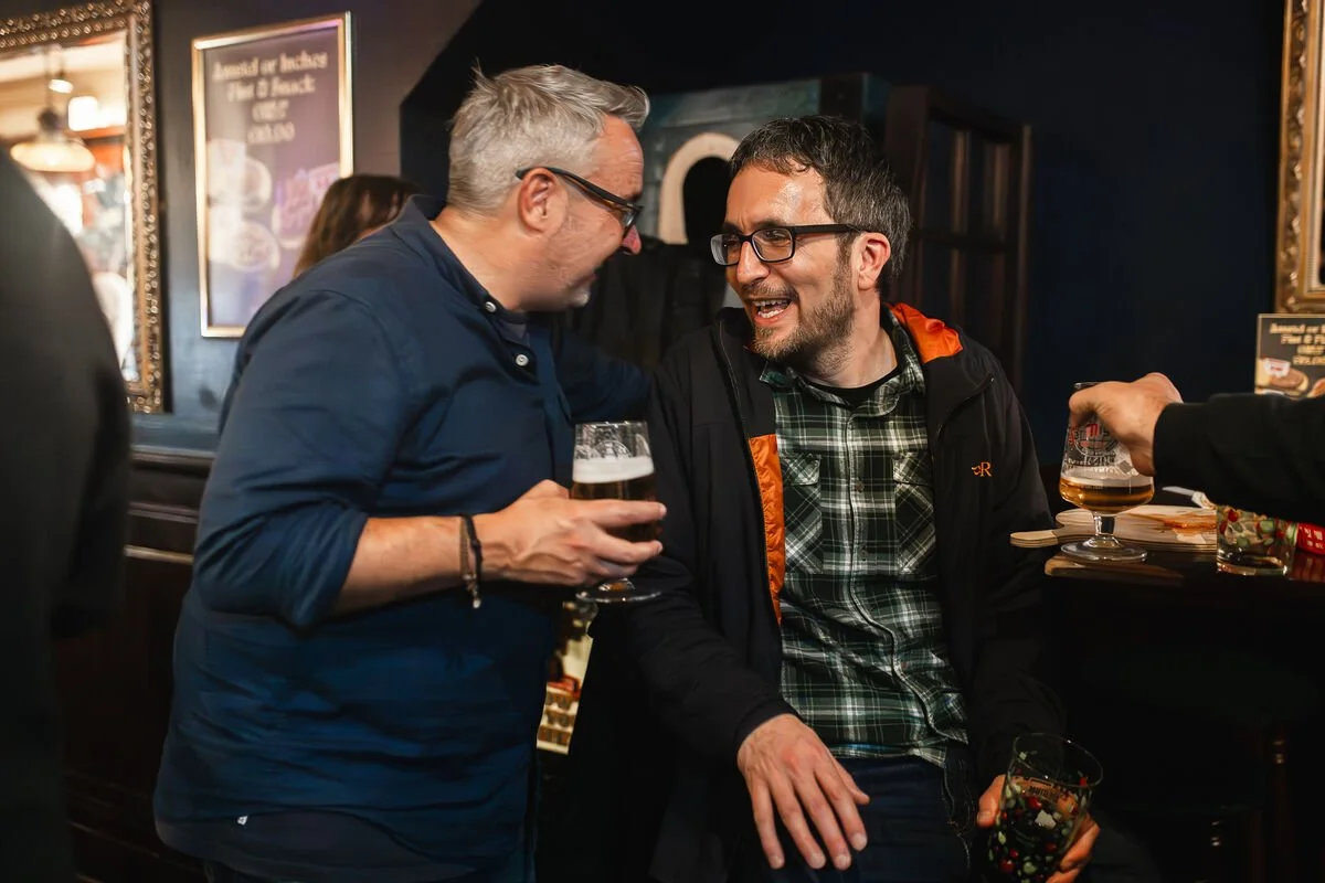 Two men are chatting and laughing at a bar, each holding a glass of beer.