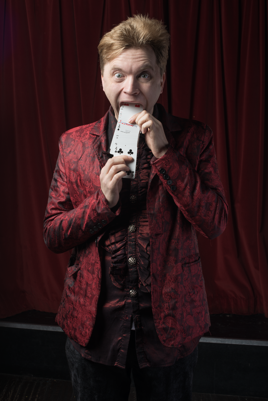 A man in a red patterned jacket shuffling cards in front of a red curtain.