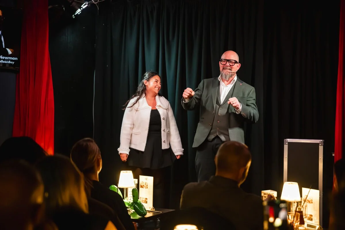 Stand-up comedy performance with a man in a green blazer and glasses, and a woman in a black dress and white jacket, on a stage with a black curtain backdrop, lit by small table lamps, in front of an audience.