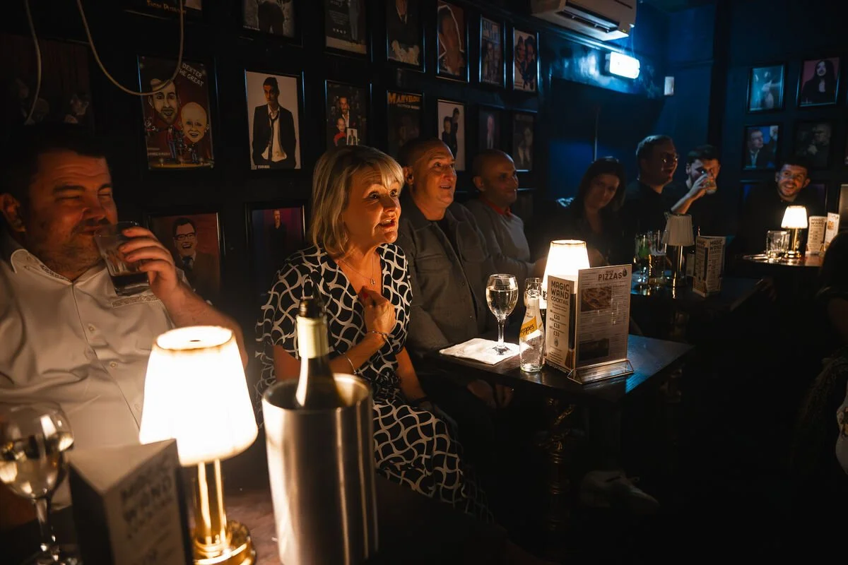 A group of people sitting at tables in a dimly lit bar or club, some with drinks, watching a performance or speaking. The walls are decorated with framed photos or artwork, and small lamps provide cozy lighting.