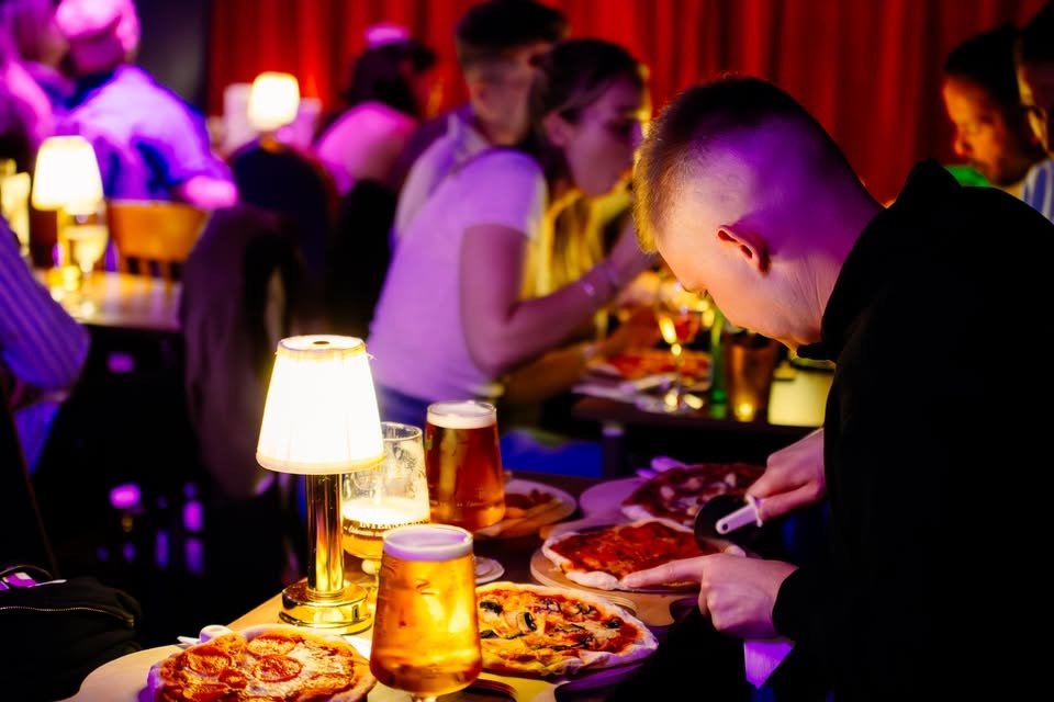 People dining at a bar with pizza and drinks, dim lighting, and colorful lights.