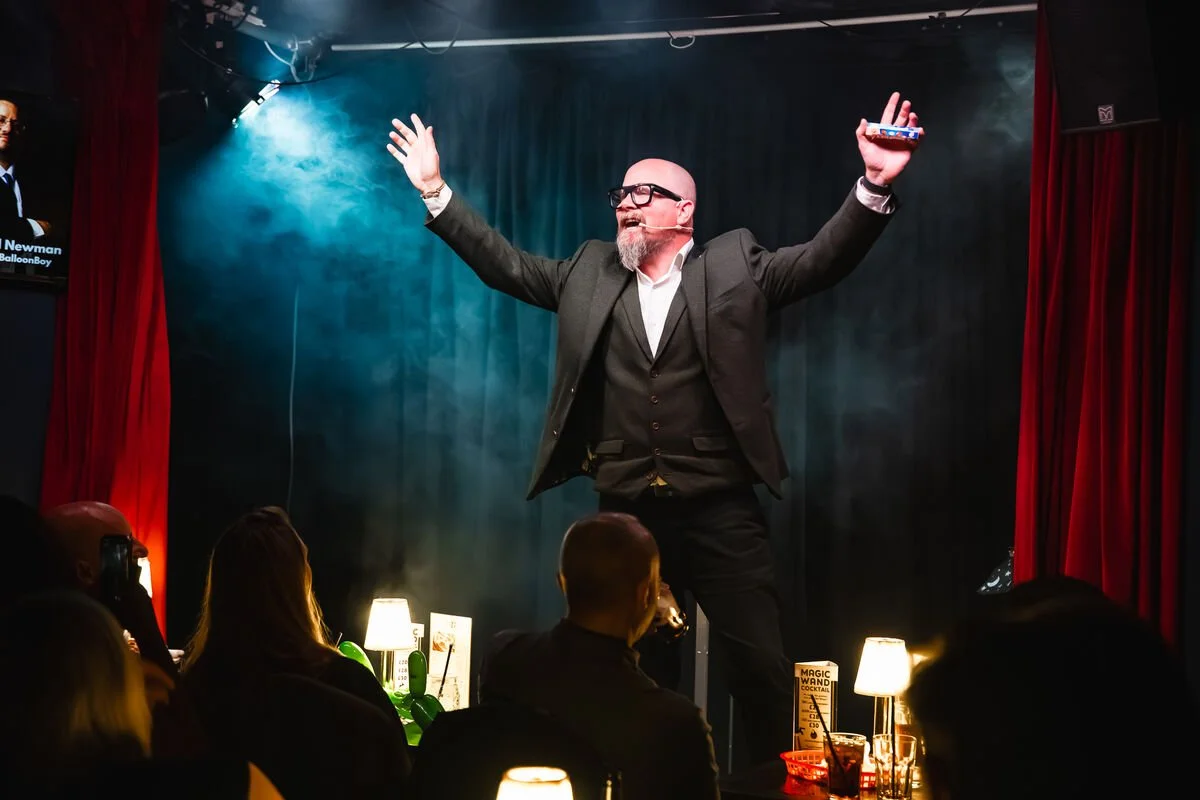 Comedian performing on stage with a microphone headset, in a dimly lit comedy club with red curtains, an audience watching.