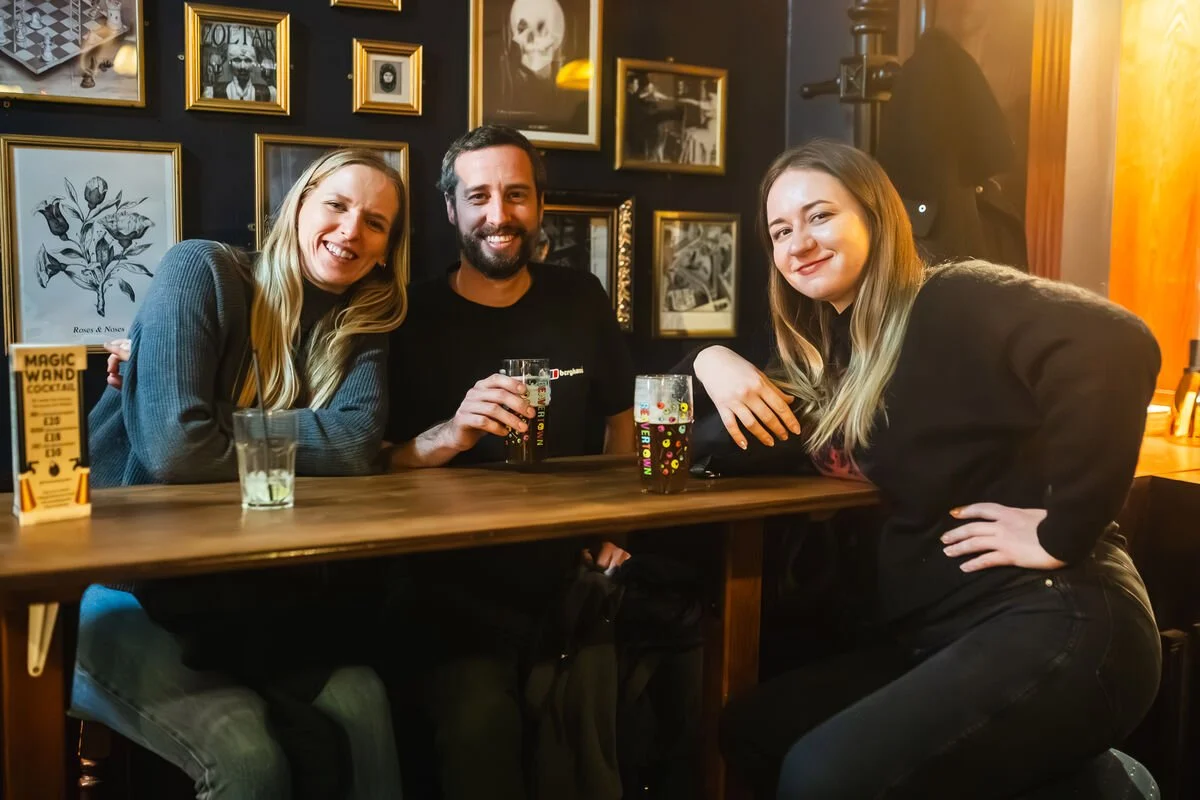 Three people sitting at a bar table drinking colorful cocktails, smiling at the camera, with decorated walls featuring framed artwork and photographs behind them.