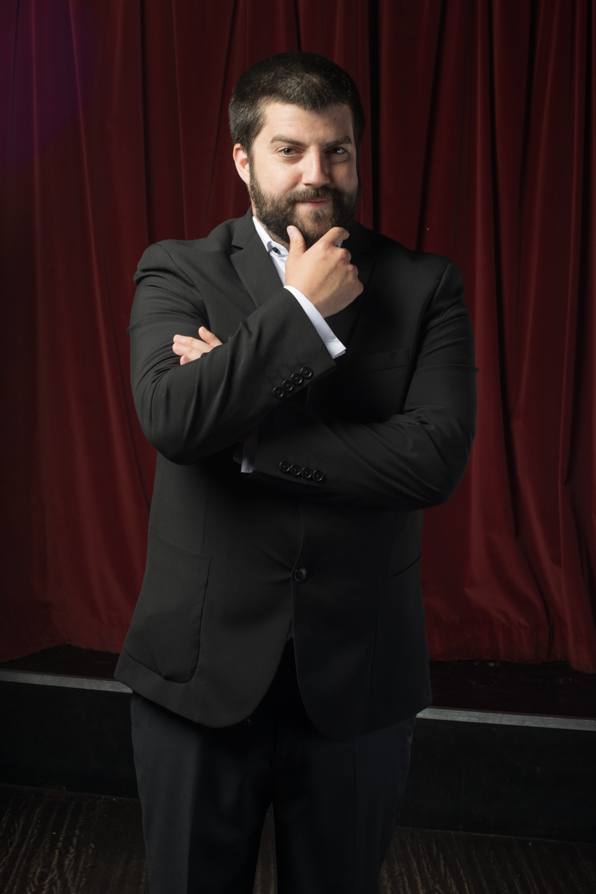 A man in a black tuxedo with a white shirt standing in front of red velvet curtains, touching his chin with one hand and crossing his arms.