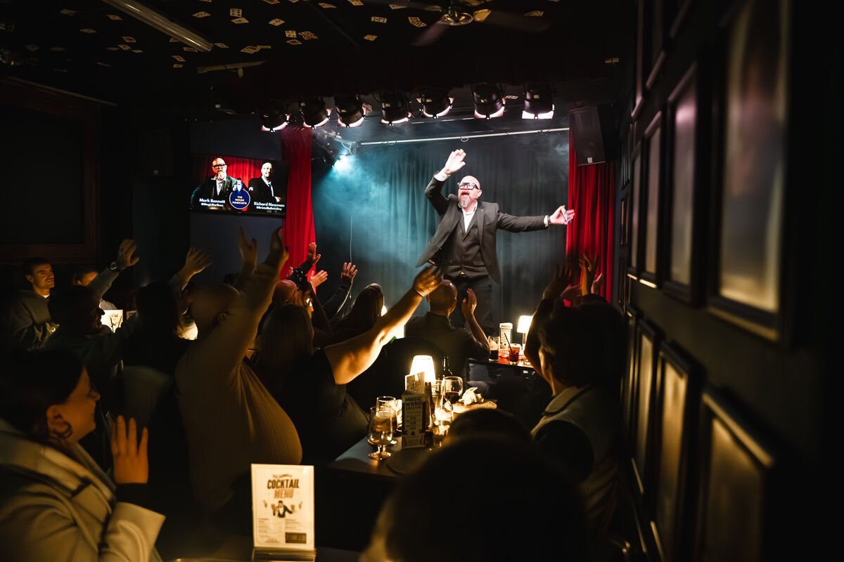 A performer on stage with a microphone, gesturing with one hand, in front of an audience raising their hands in a dimly lit comedy club or theater.