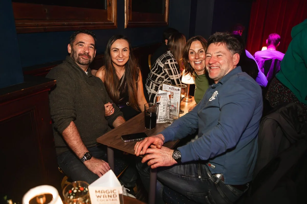 Four people sitting at a bar or restaurant table smiling for the camera, with drinks in front of them and a menu on the table.