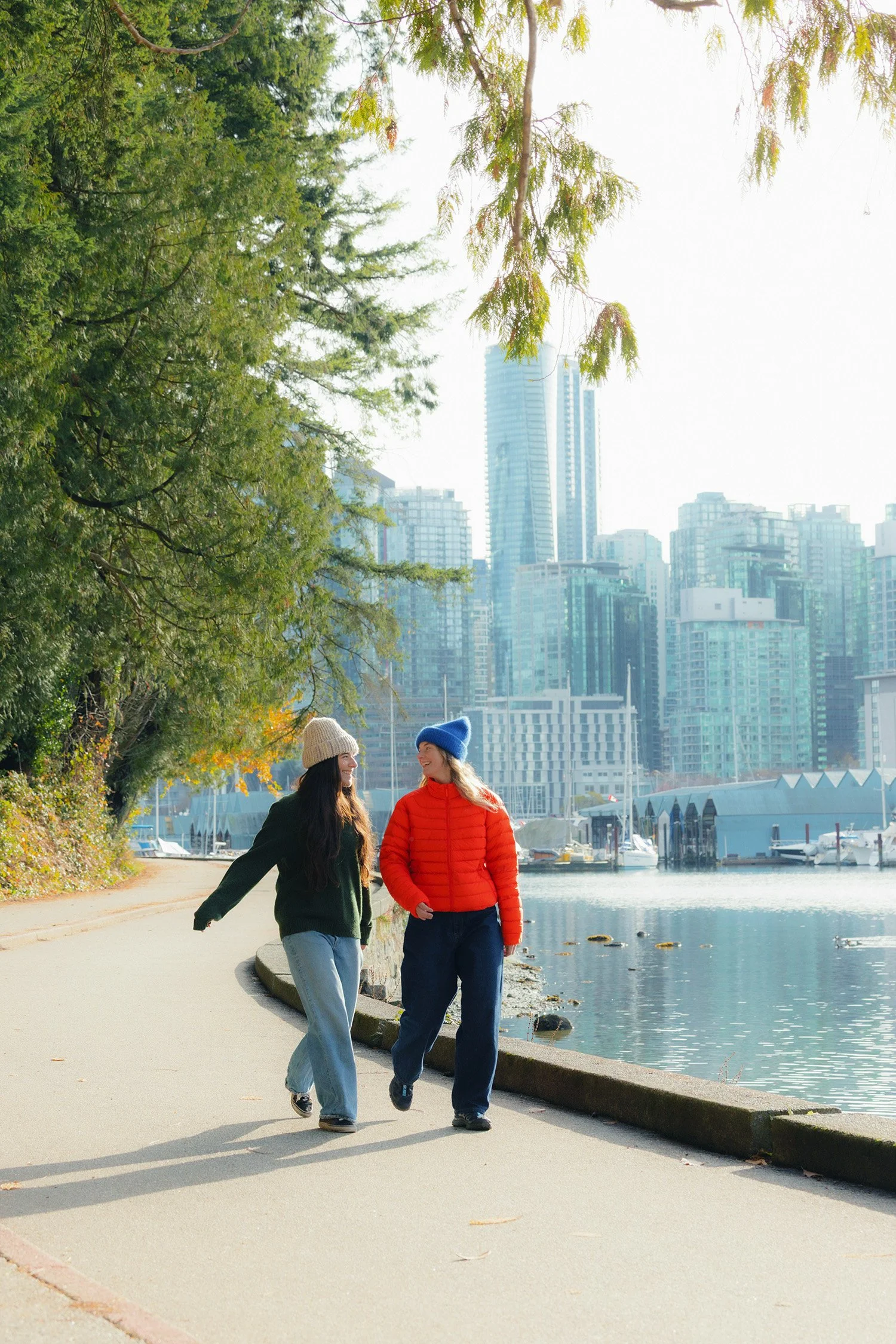 Two youth girls walking on a pedestrian path next to the water, with the backdrop of Vancouver city buildings and trees