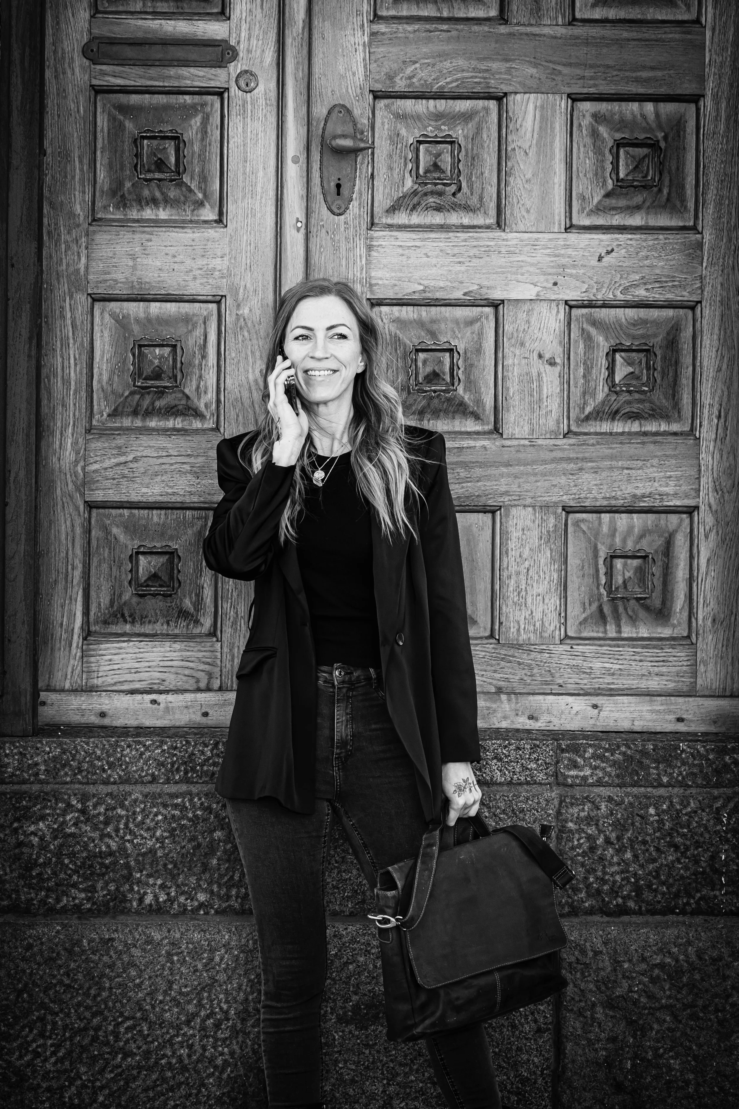 A woman standing in front of a wooden door, smiling while talking on her cellphone, holding a bag, and wearing a black blazer and jeans.