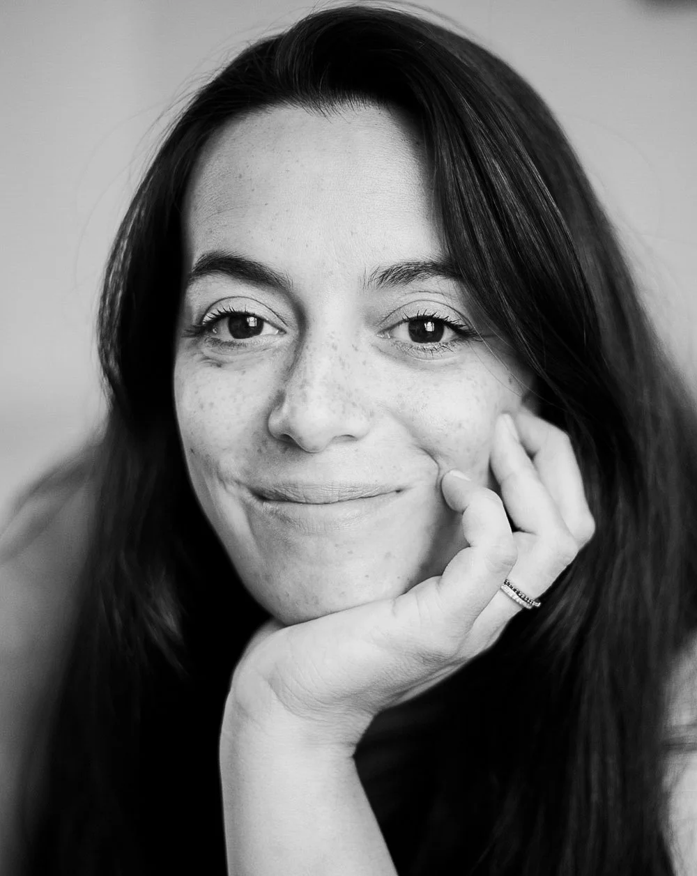 Black and white close-up portrait of a woman with long dark hair, smiling softly, resting her chin on her hand, and wearing a ring on her finger.