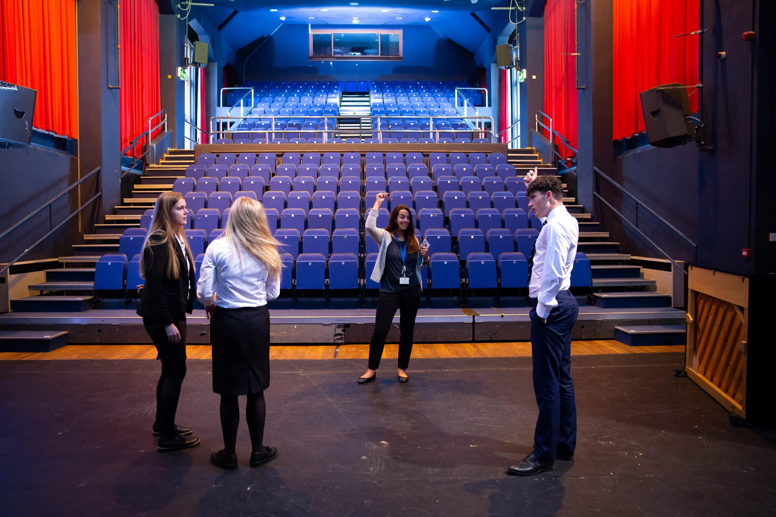Four people standing on a stage inside a theater, with a woman in the middle raising her fist, and four empty blue seats behind them.