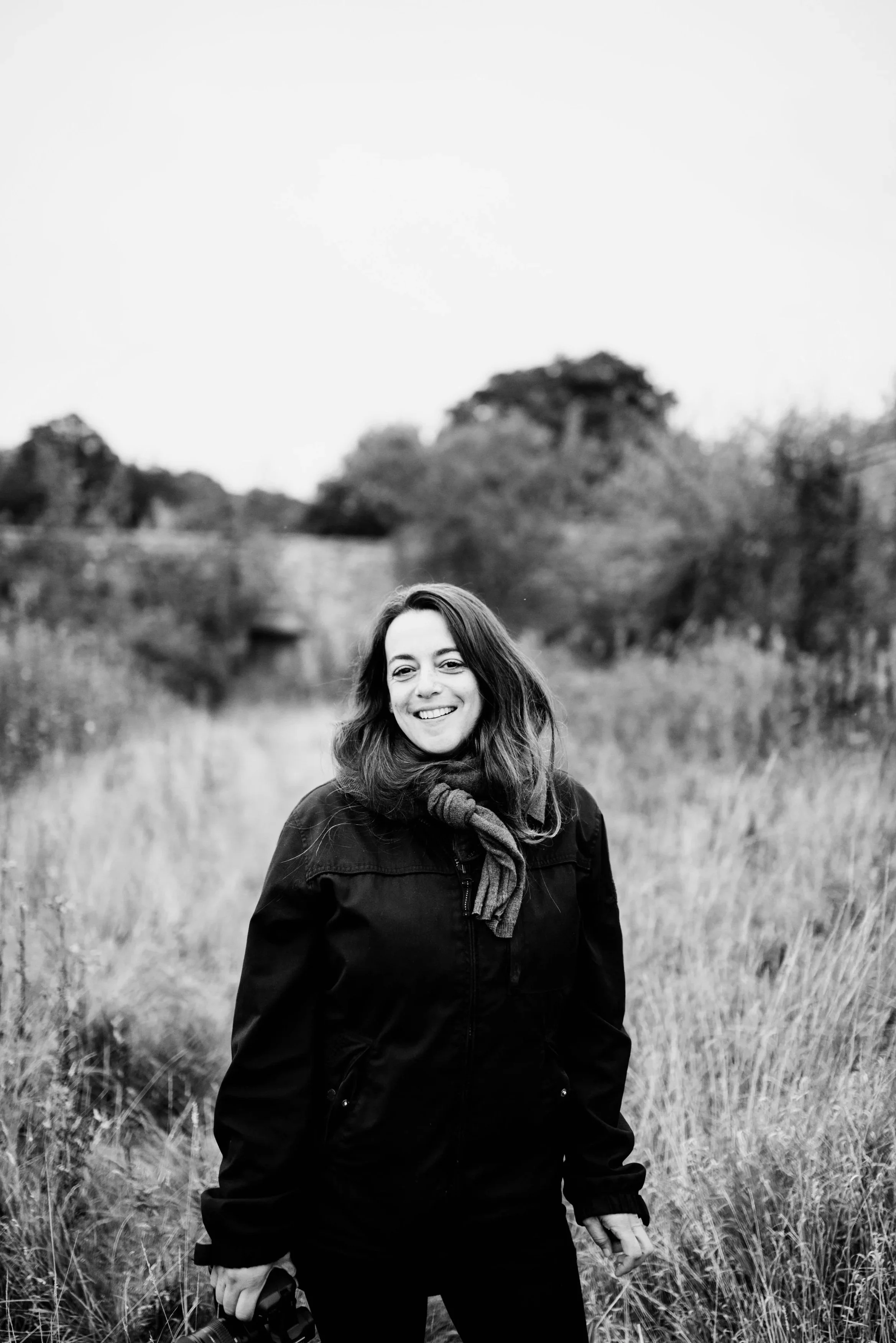 Black and white photo of a woman outdoors smiling, standing in tall grass with trees in the background.