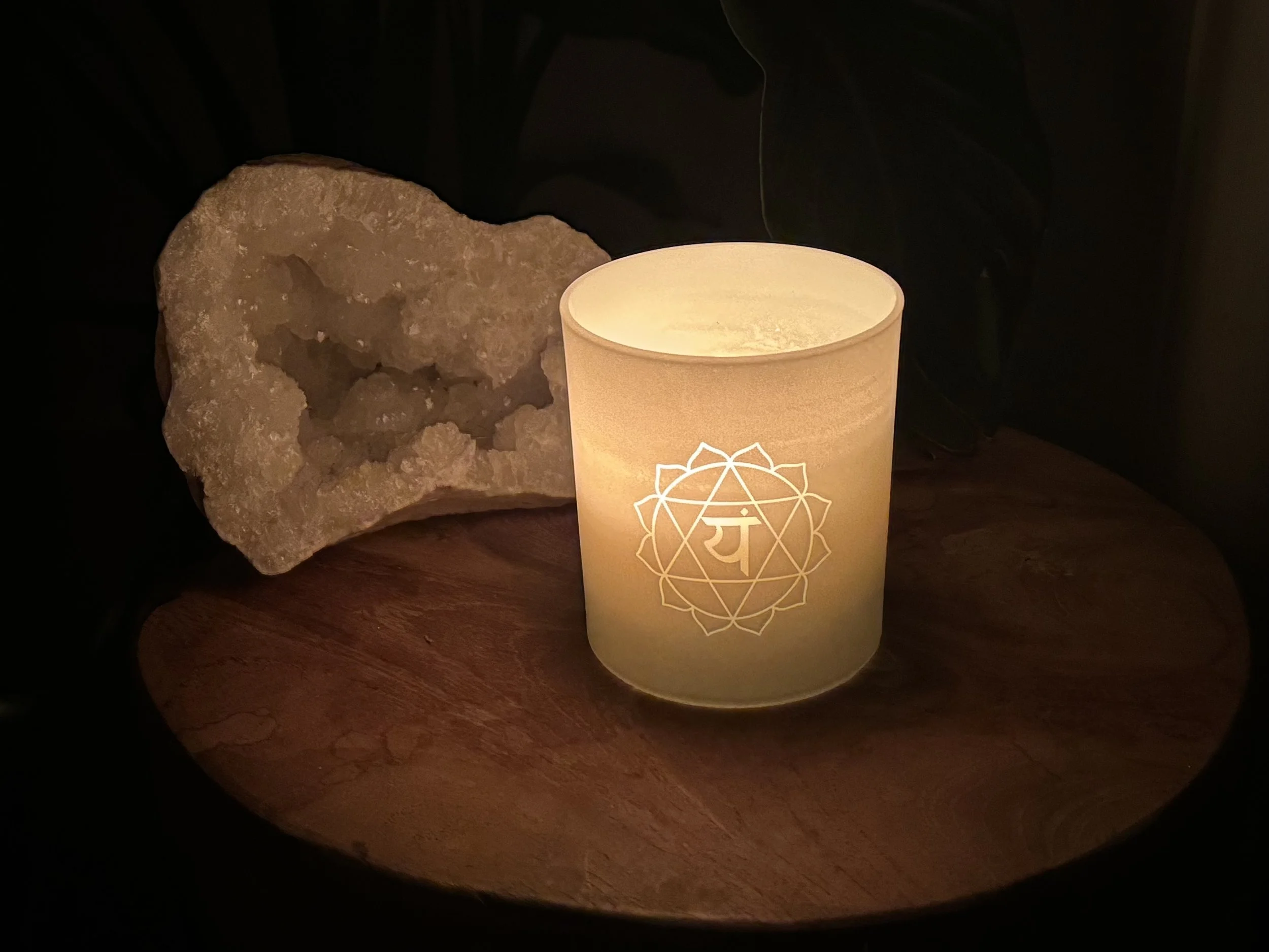 A glowing candle with a mandala design and a Hindi symbol on it, placed on a wooden surface next to a large, rough crystal in a dark setting.