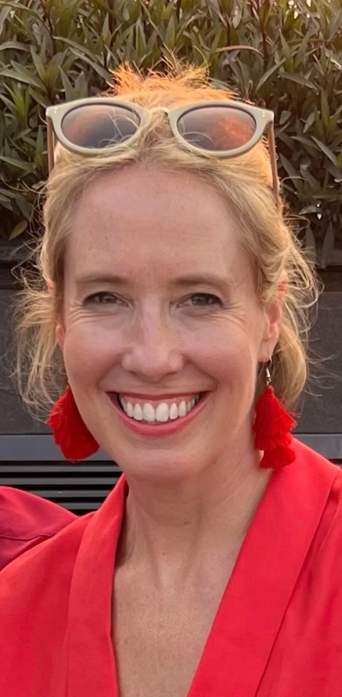 A woman smiling, wearing red earrings and a red top, with sunglasses on her head, outdoors with greenery in the background.