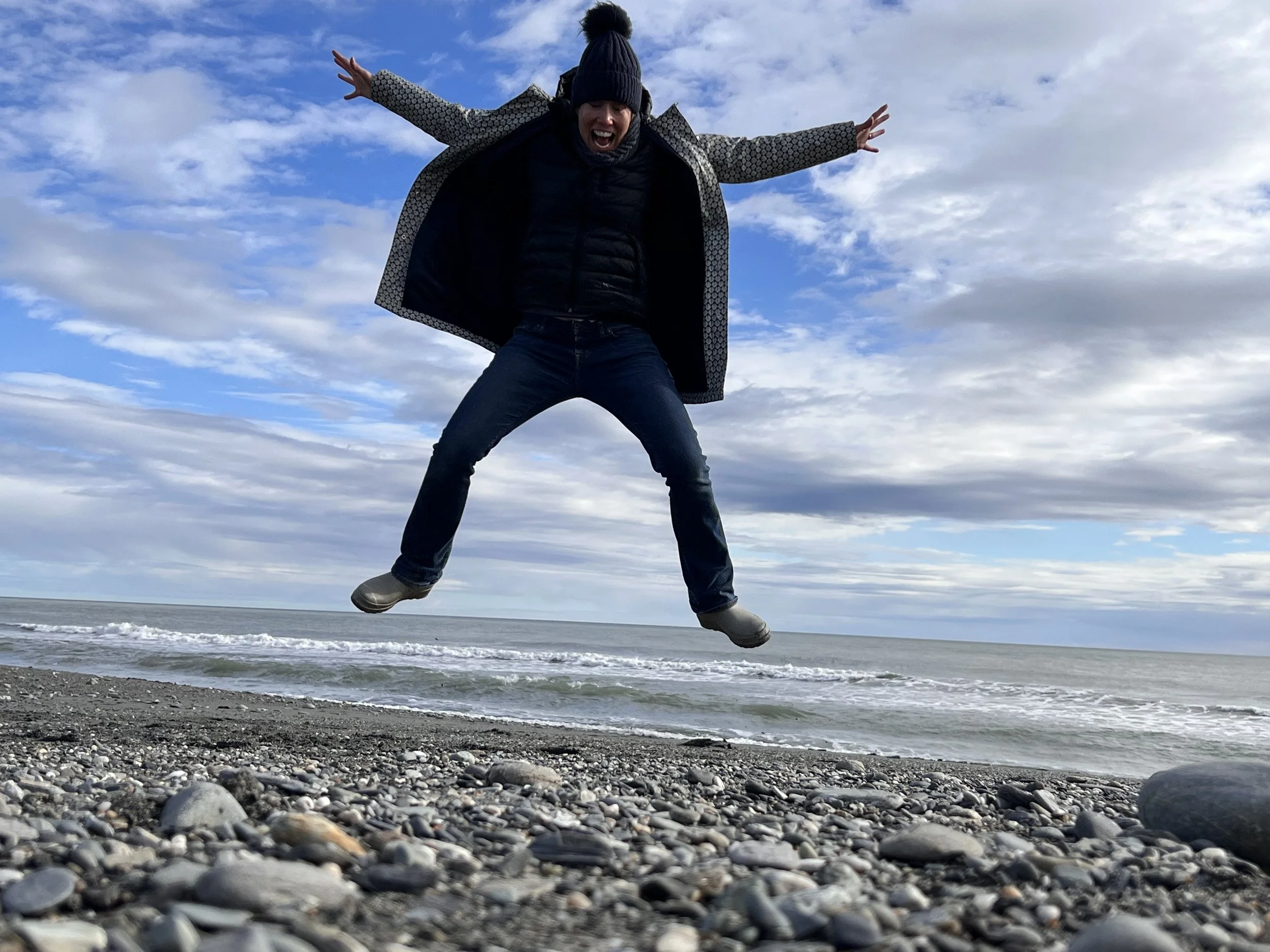 Person jumping on pebbled beach with waves and cloudy sky in the background.