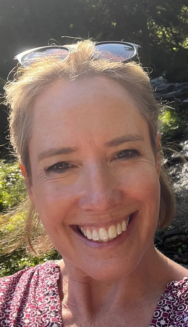 Close-up of woman smiling outdoors, with sunglasses on her head and greenery in the background.