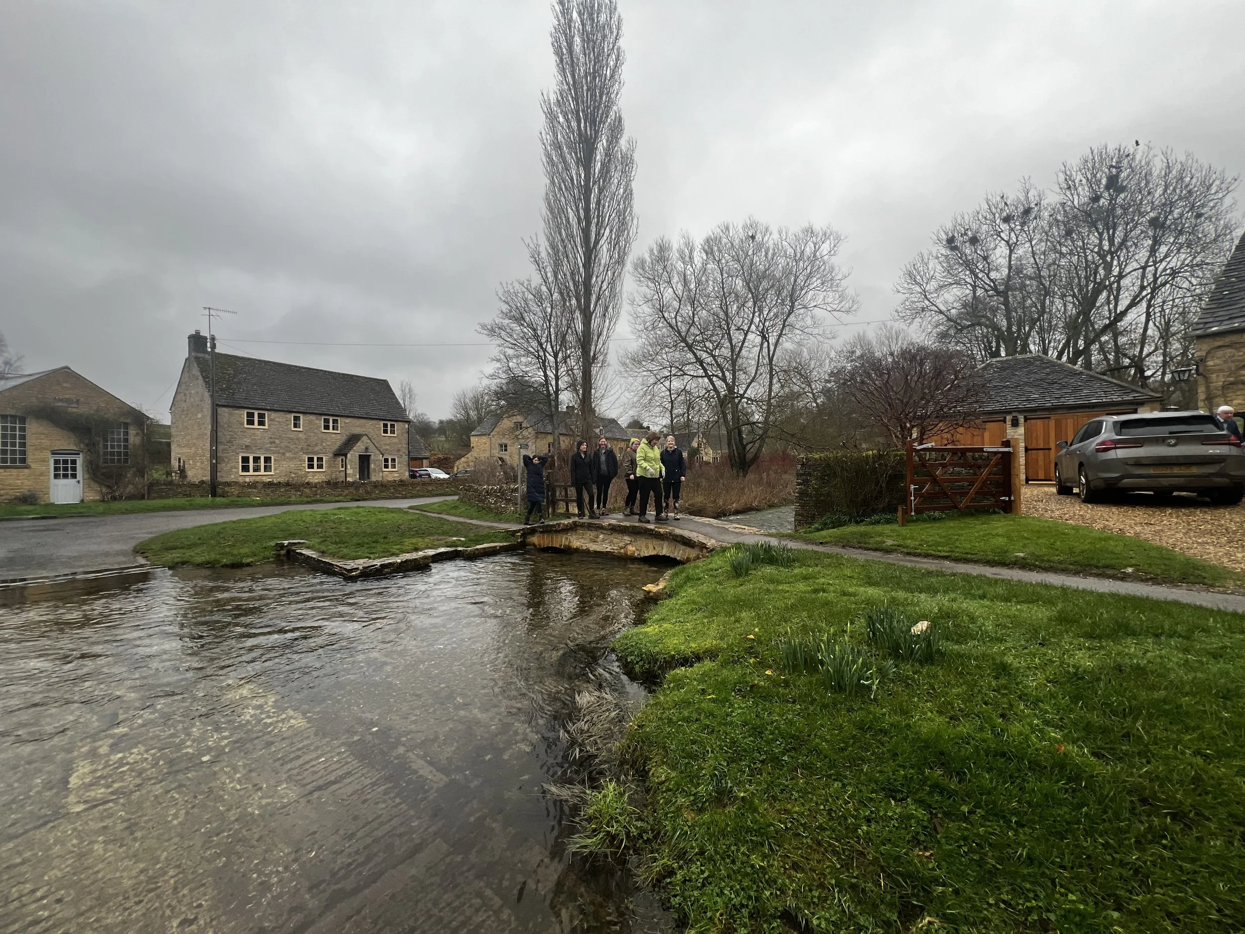 A group of people standing on a small stone bridge crossing a river in a village with stone houses and leafless trees, under a cloudy sky.