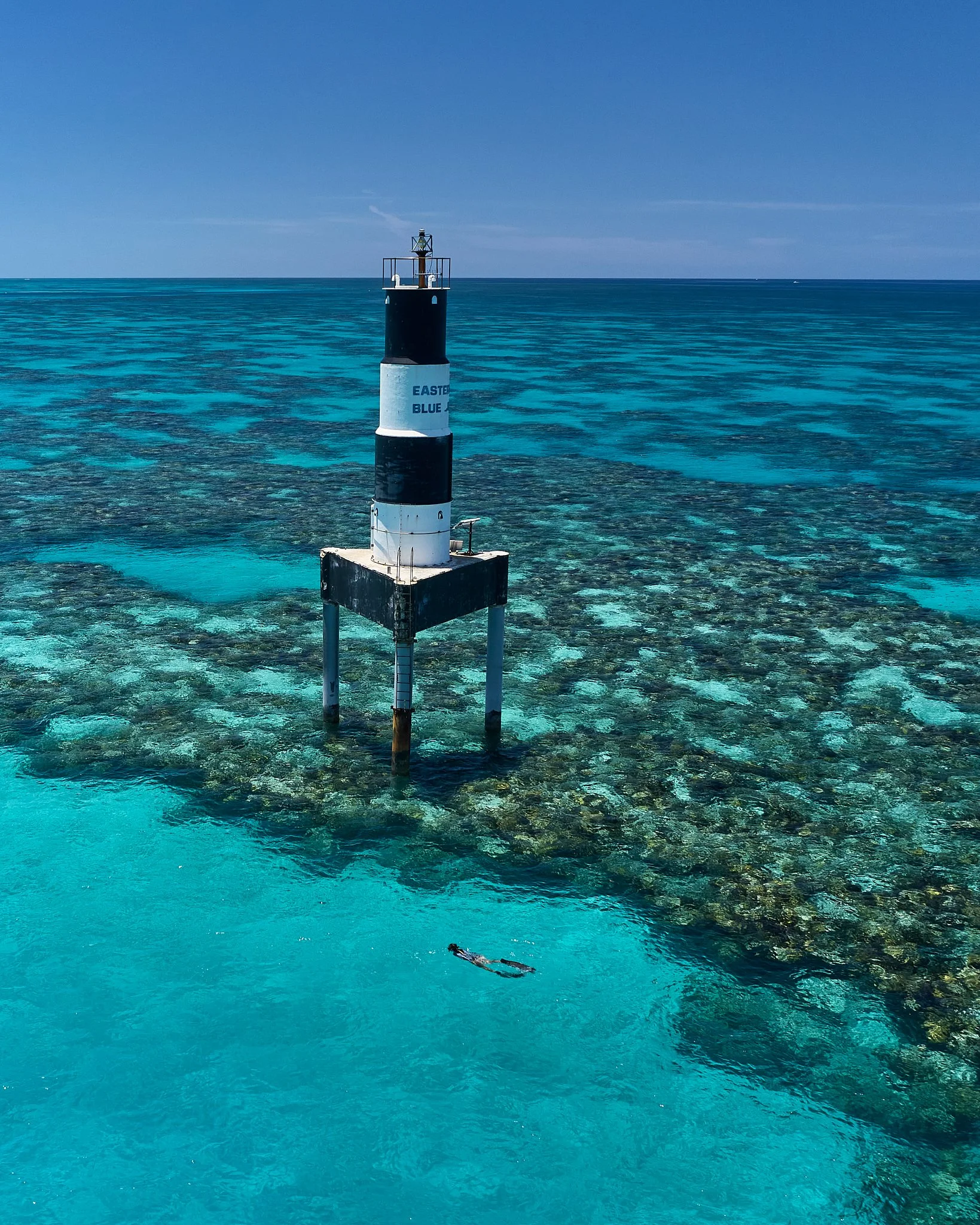A black and white lighthouse standing on stilts in the clear turquoise ocean water with coral underneath.