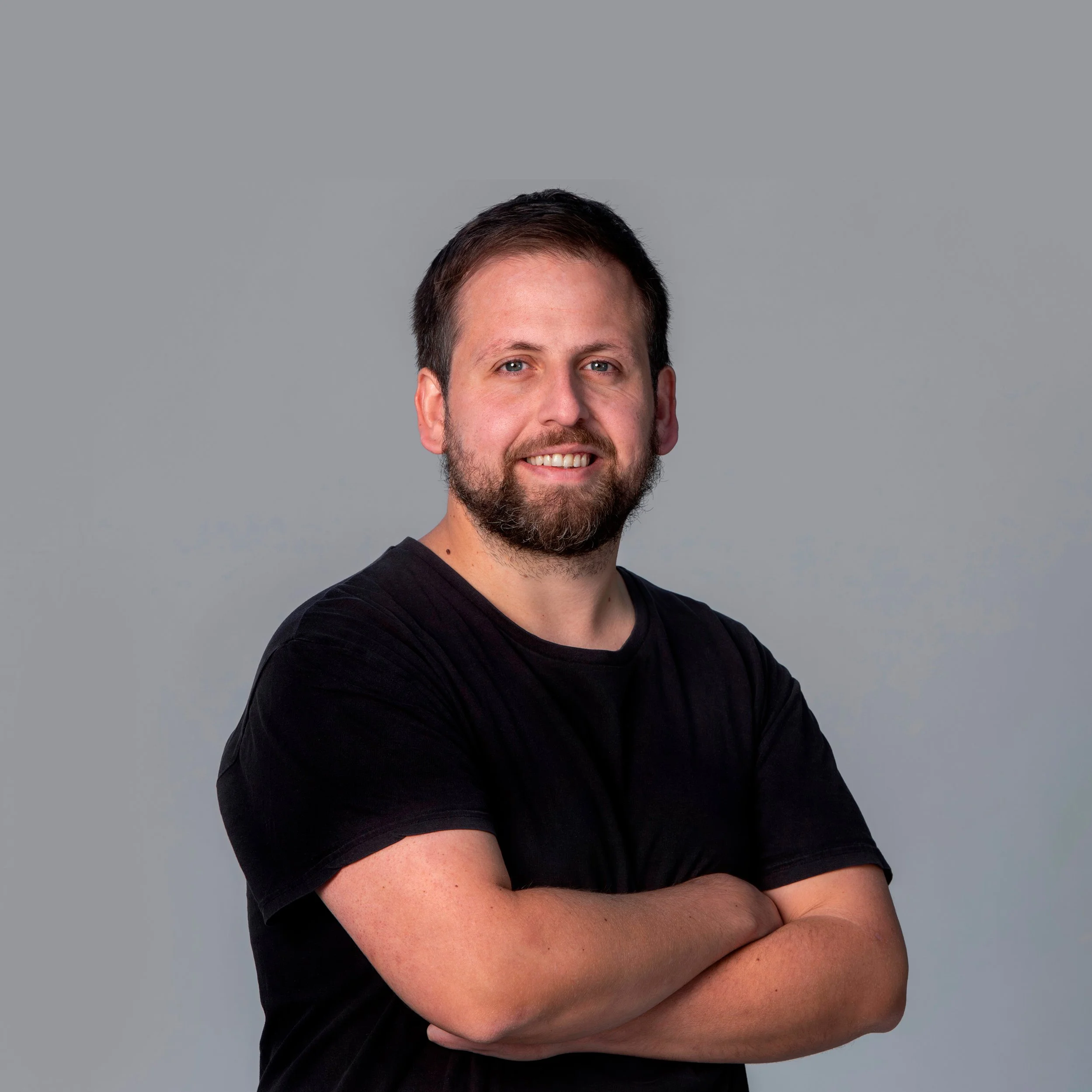 Retrato de un hombre con barba y cabello castaño oscuro, sonriendo, con brazos cruzados, puesto en un fondo de color gris claro.