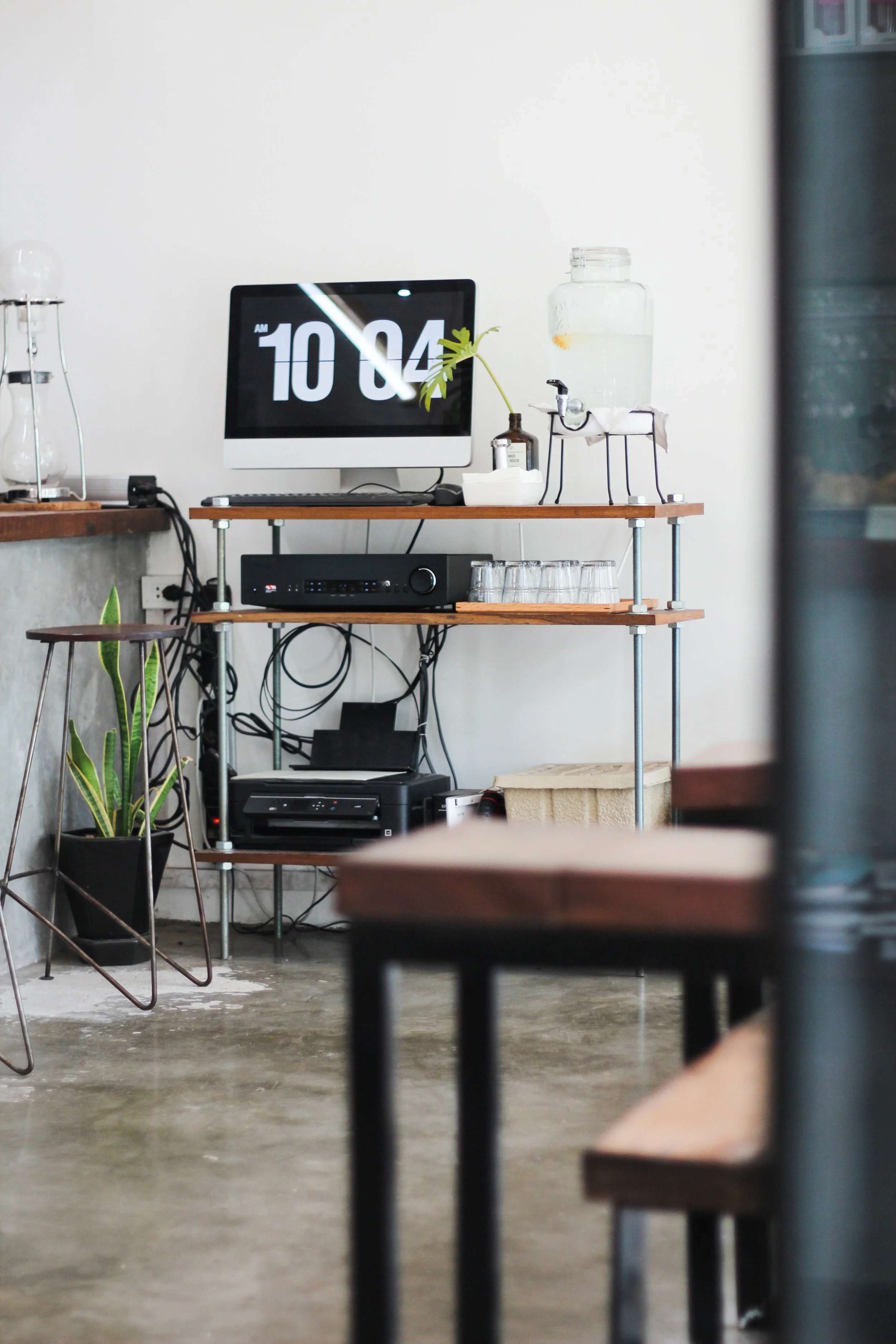Una estantería de metal y madera con un monitor, una planta, un dispensador de agua, vasos apilados y equipo de audio.