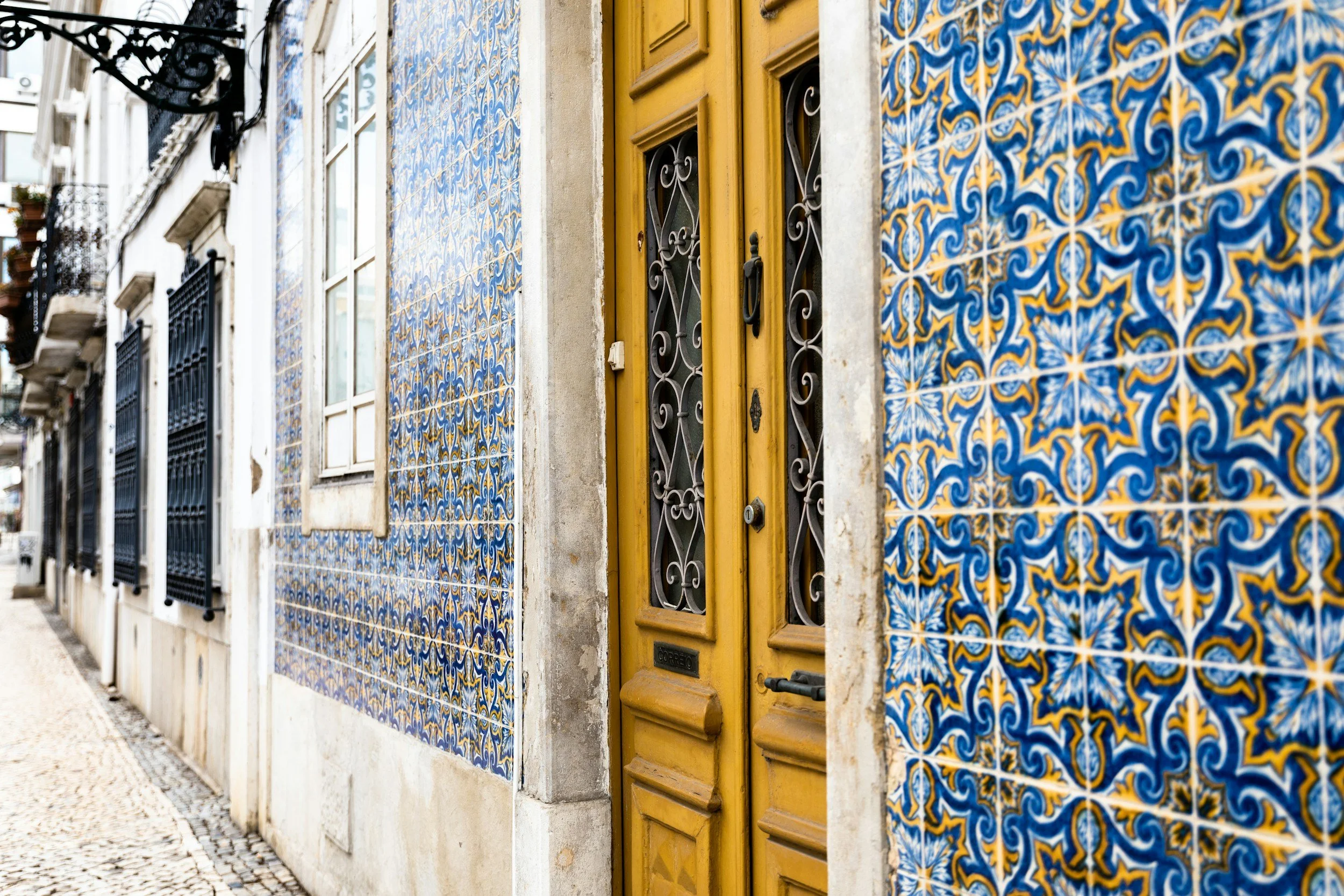 A yellow wooden door with black wrought iron detailing, set in a building with decorative blue and yellow patterned tiles on the facade. The street has cobblestones and the building features black metal window guards.