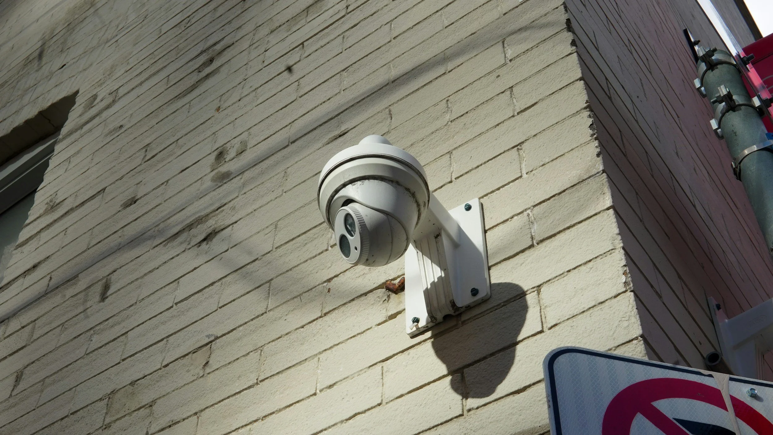 Outdoor security camera mounted on a beige brick wall, with a no parking sign nearby.