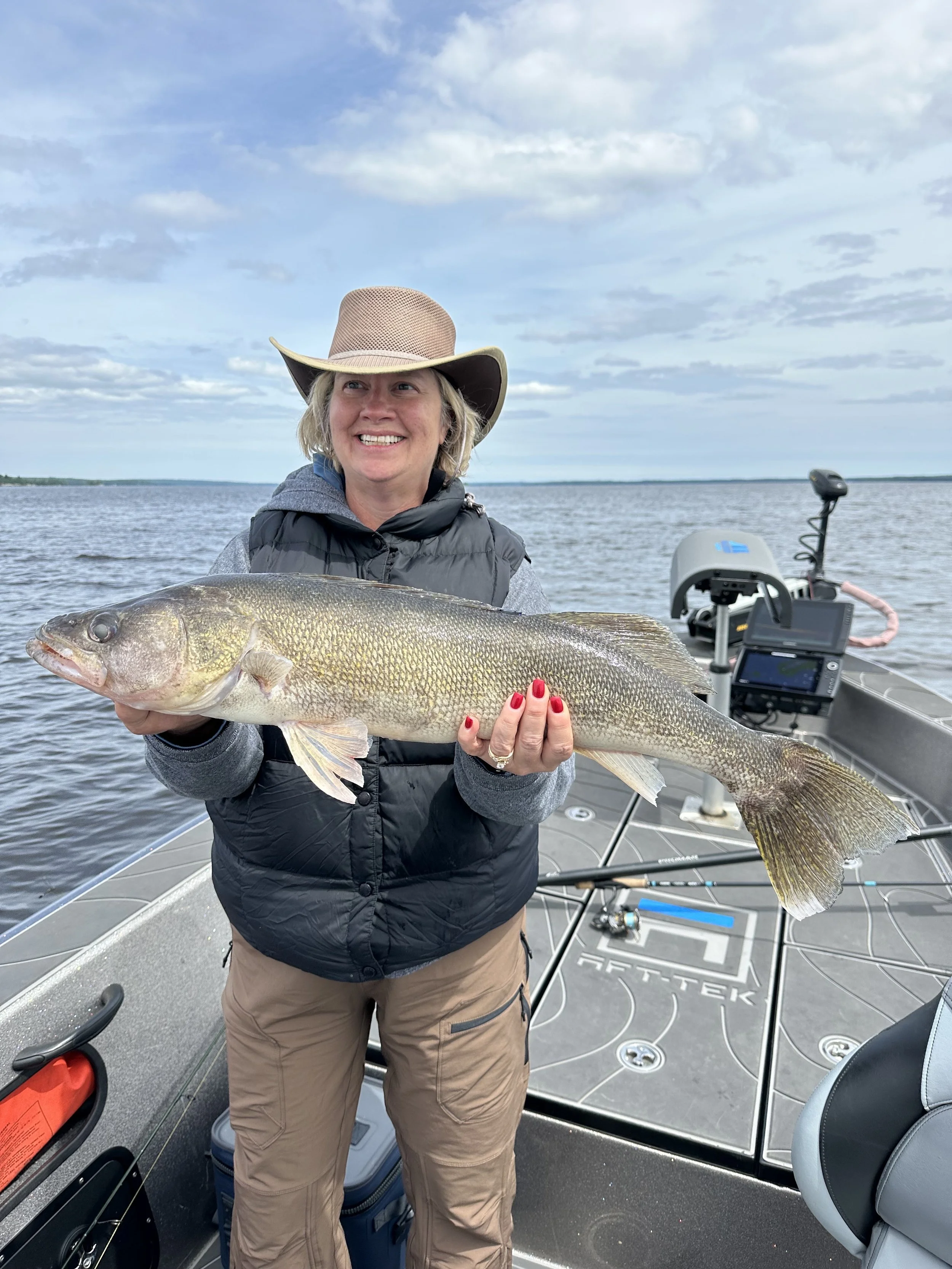 A woman on a boat holding a large fish she caught, smiling with an overcast sky and water in the background.