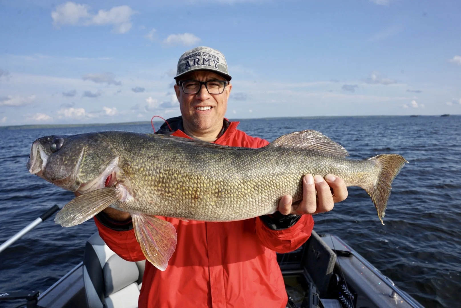A man wearing glasses, a gray cap with 'United States Army' on it, and a red jacket, holding a large fish aboard a boat on the water. The sky is blue with some clouds.
