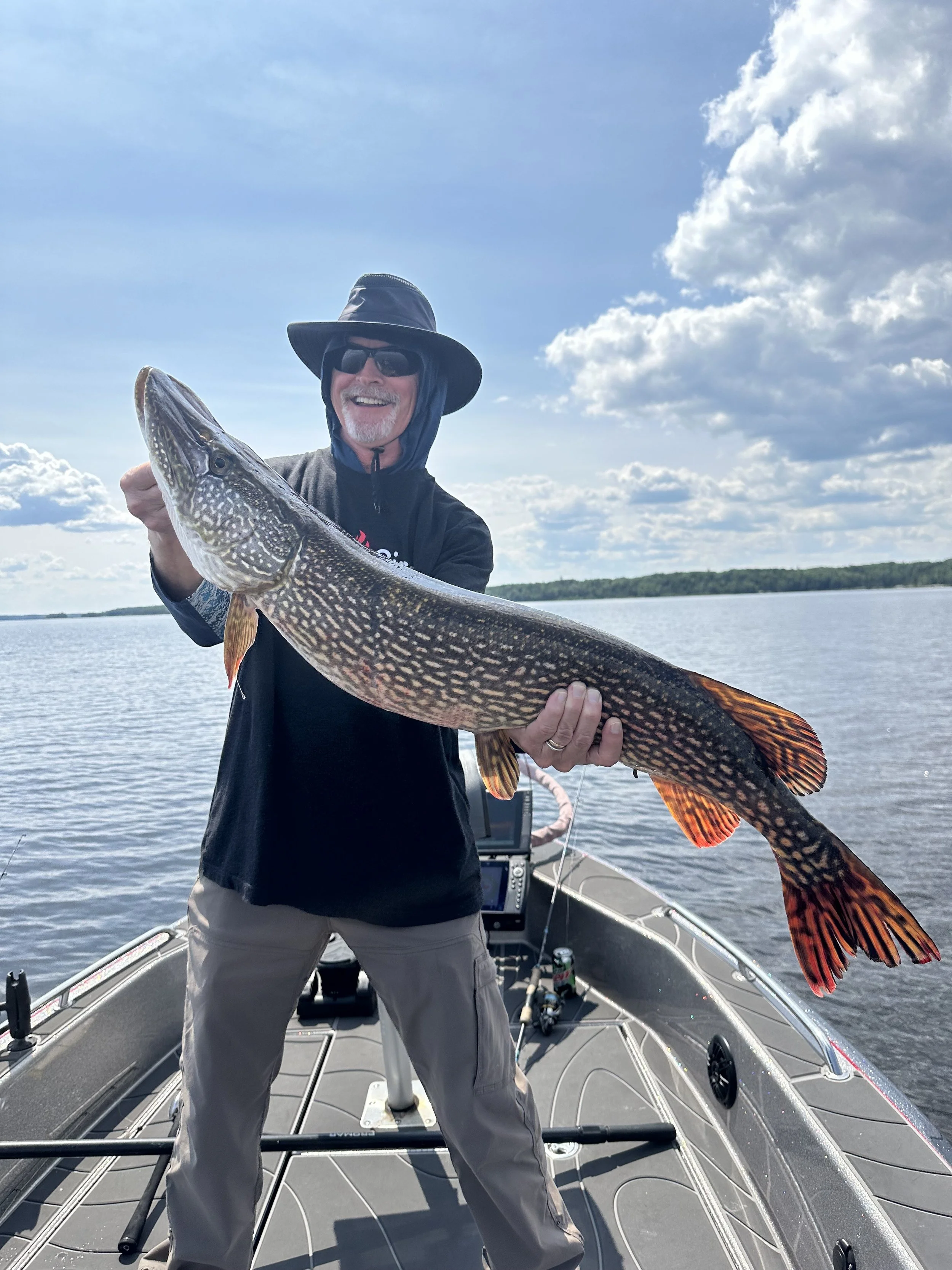 Man standing on boat holding a large fish, lake and cloudy sky in background.