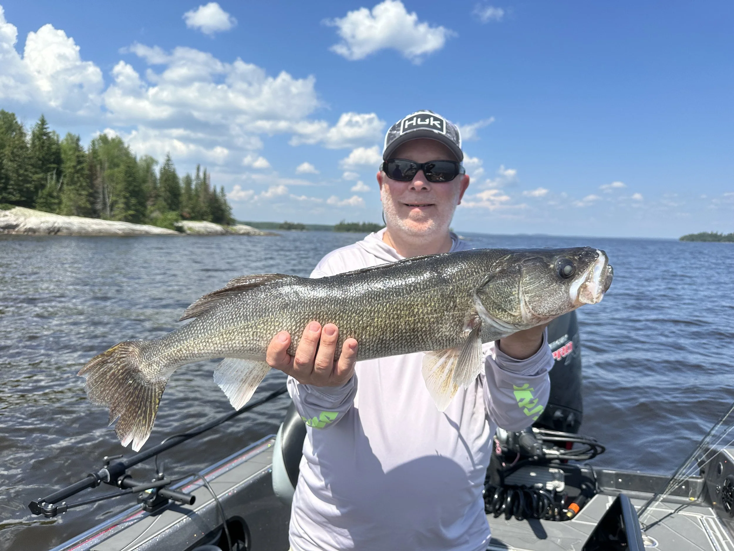 A man wearing sunglasses and a gray hoodie holding a large fish on a boat in a lake with a tree-lined shoreline under a partly cloudy sky.
