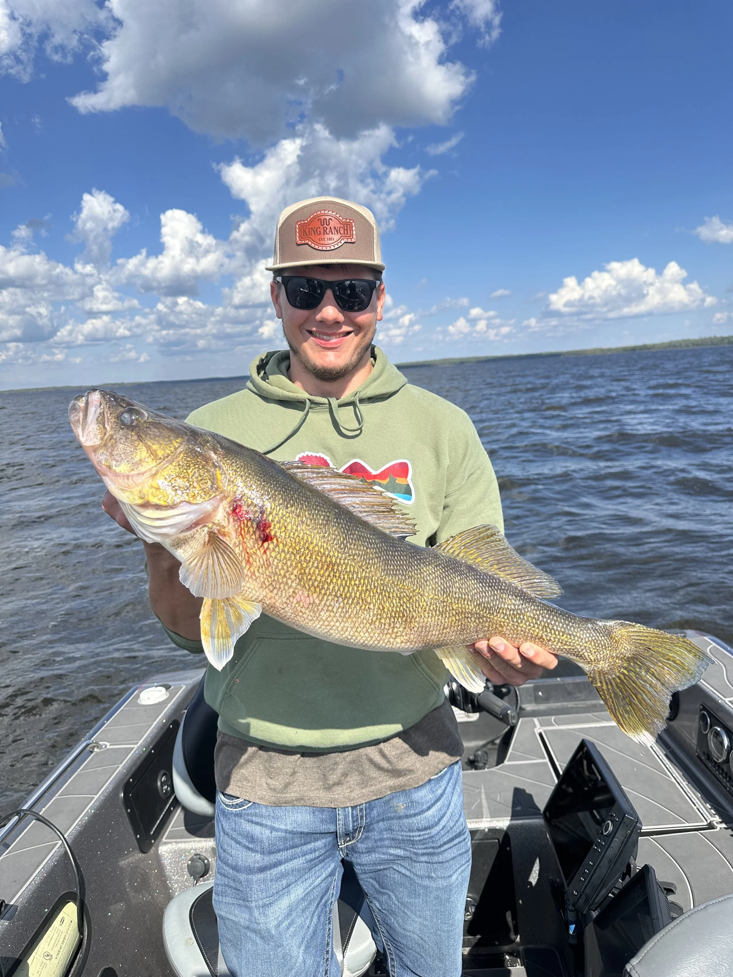 A man in sunglasses and a hoodie holding a large fish on a boat with water and sky in the background.
