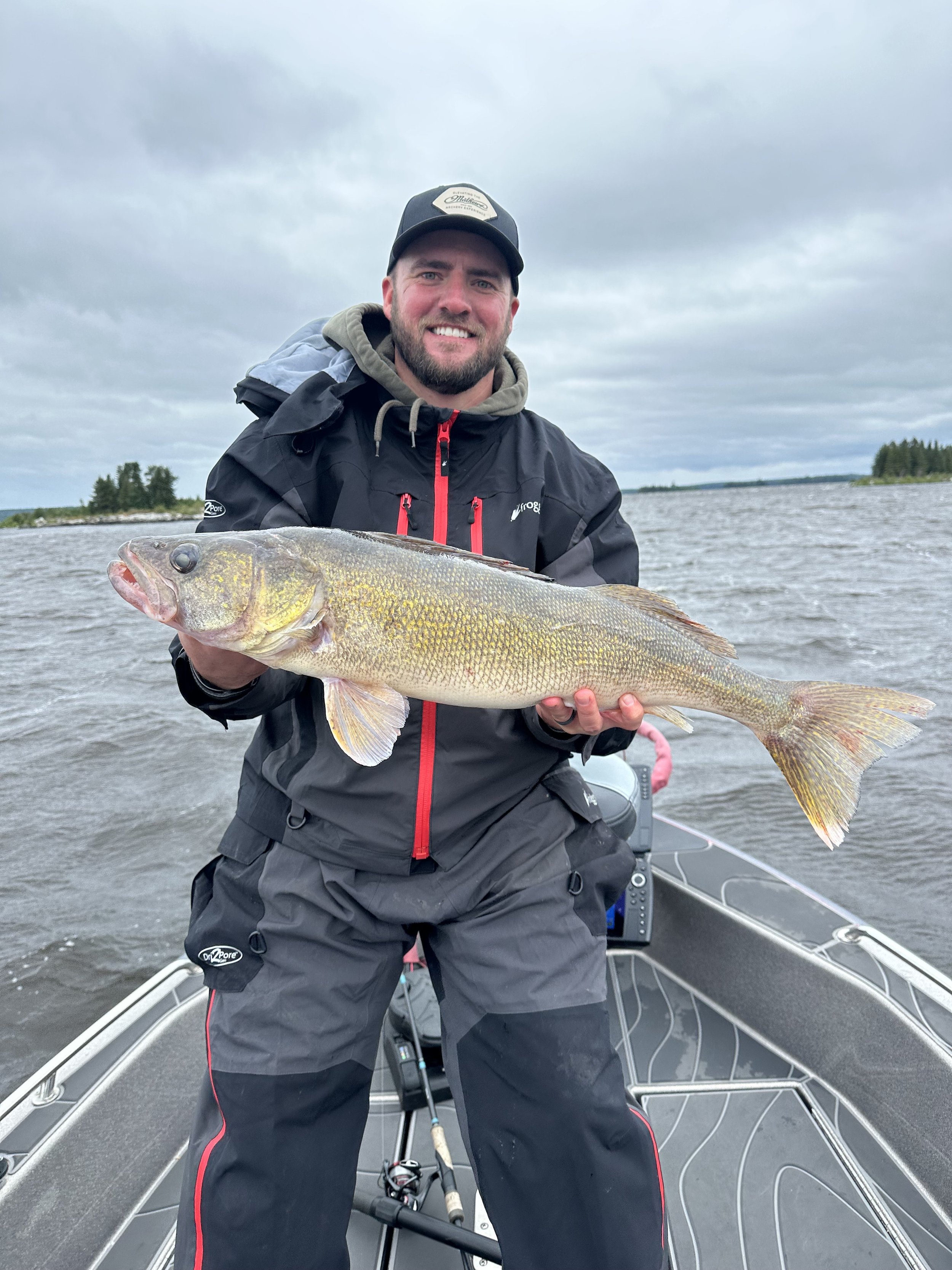 A man smiling on a boat holding a large fish, with a body of water and cloudy sky in the background.