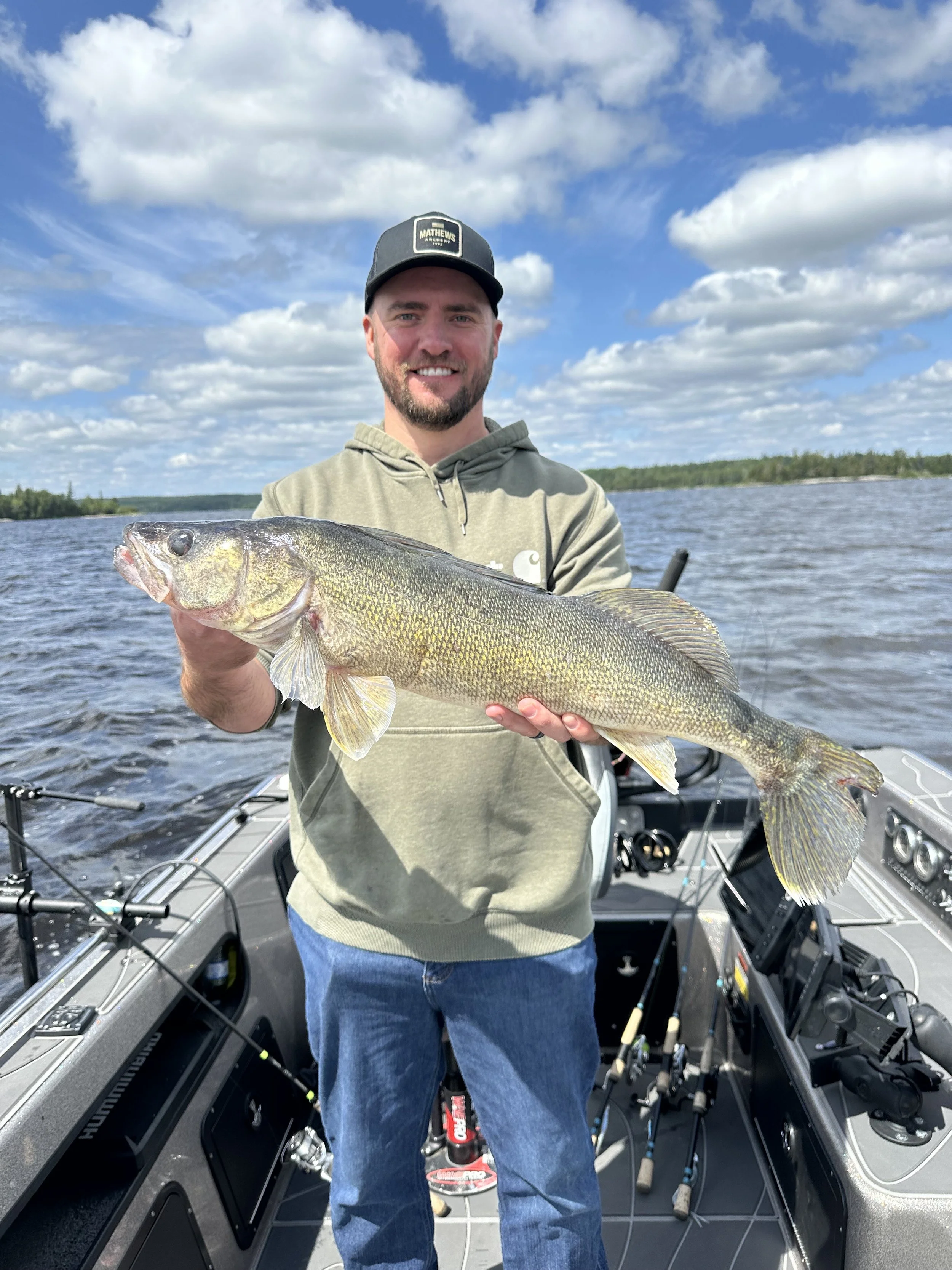 A man smiling on a boat holding a large fish he caught, with a lake and blue sky with clouds in the background.